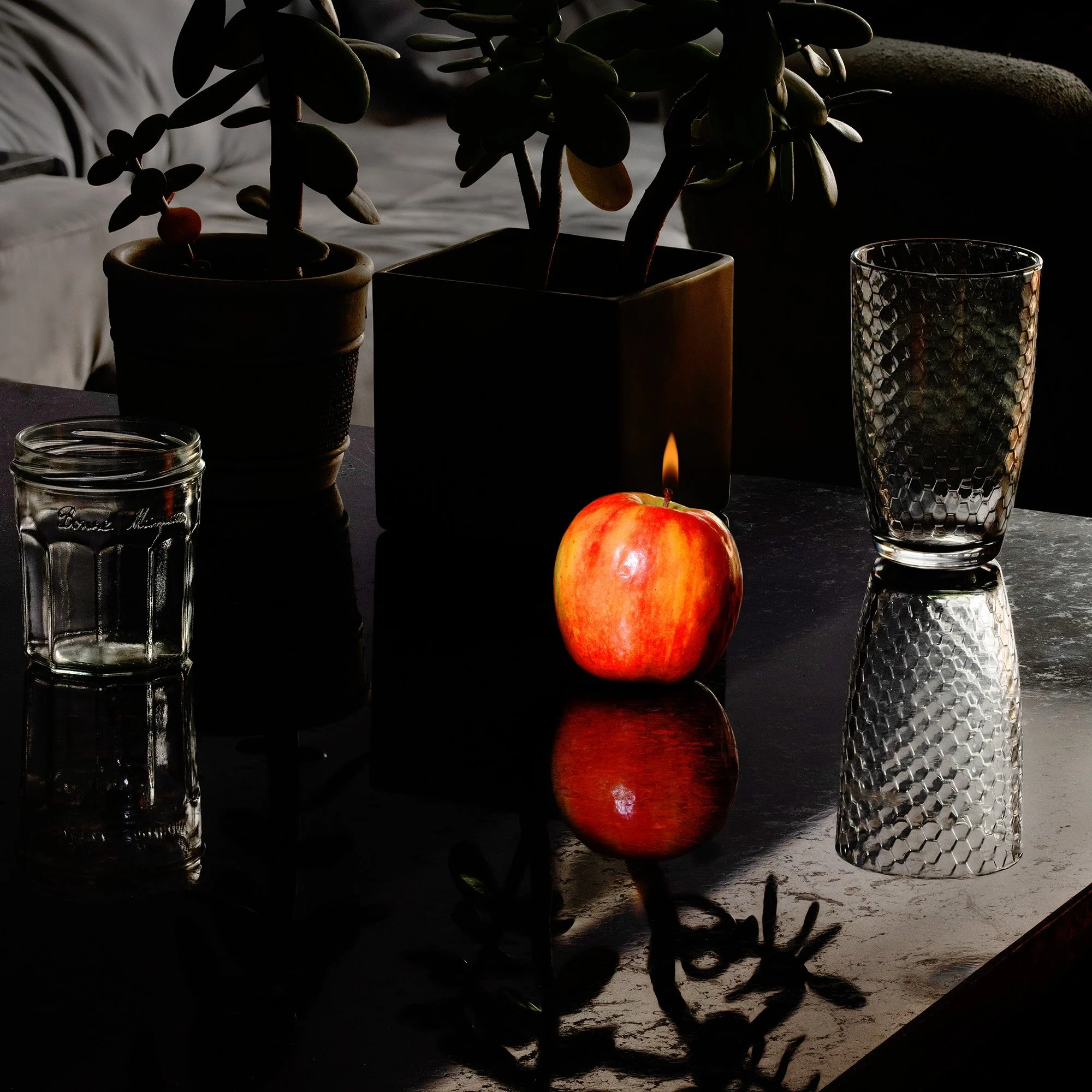 A table with a lit red apple, a candle, two empty glasses, a potted plant, and a dark background.