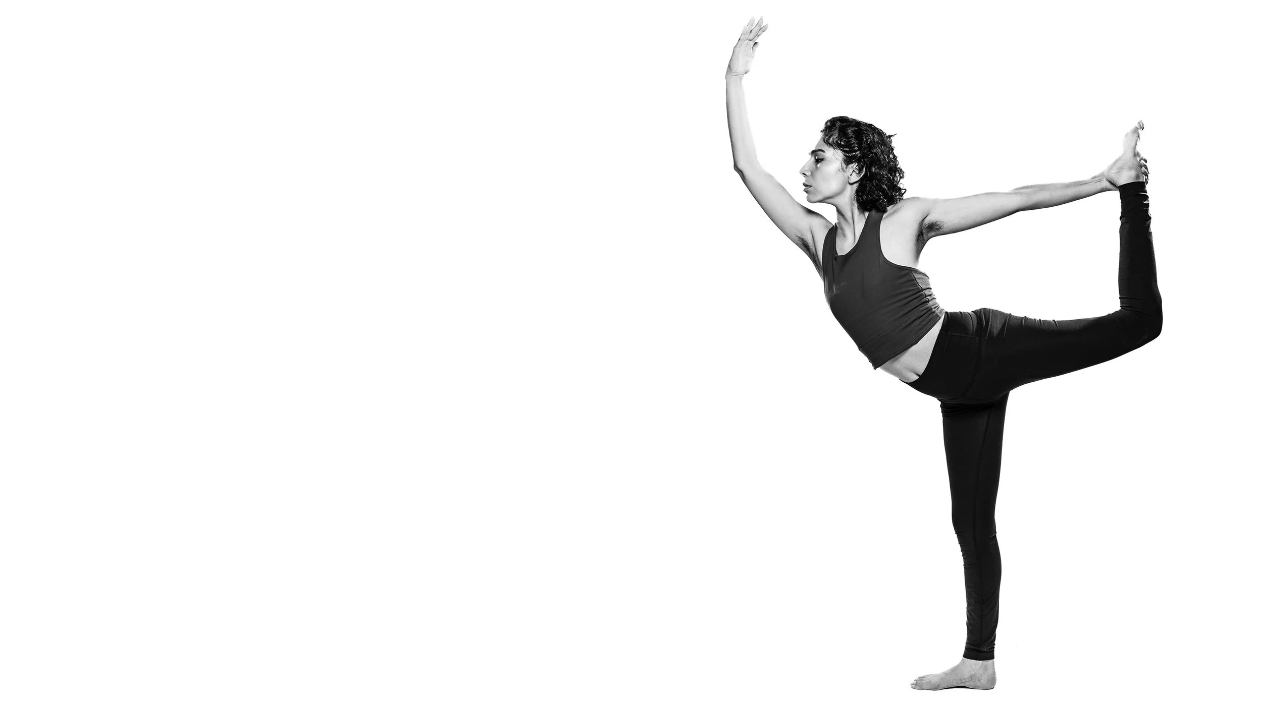 A woman with curly hair doing a yoga pose called Natarajasana or Lord of the Dance pose, balancing on one foot with the other leg bent behind her and held by her hand, while extending her other arm forward, in black and white.