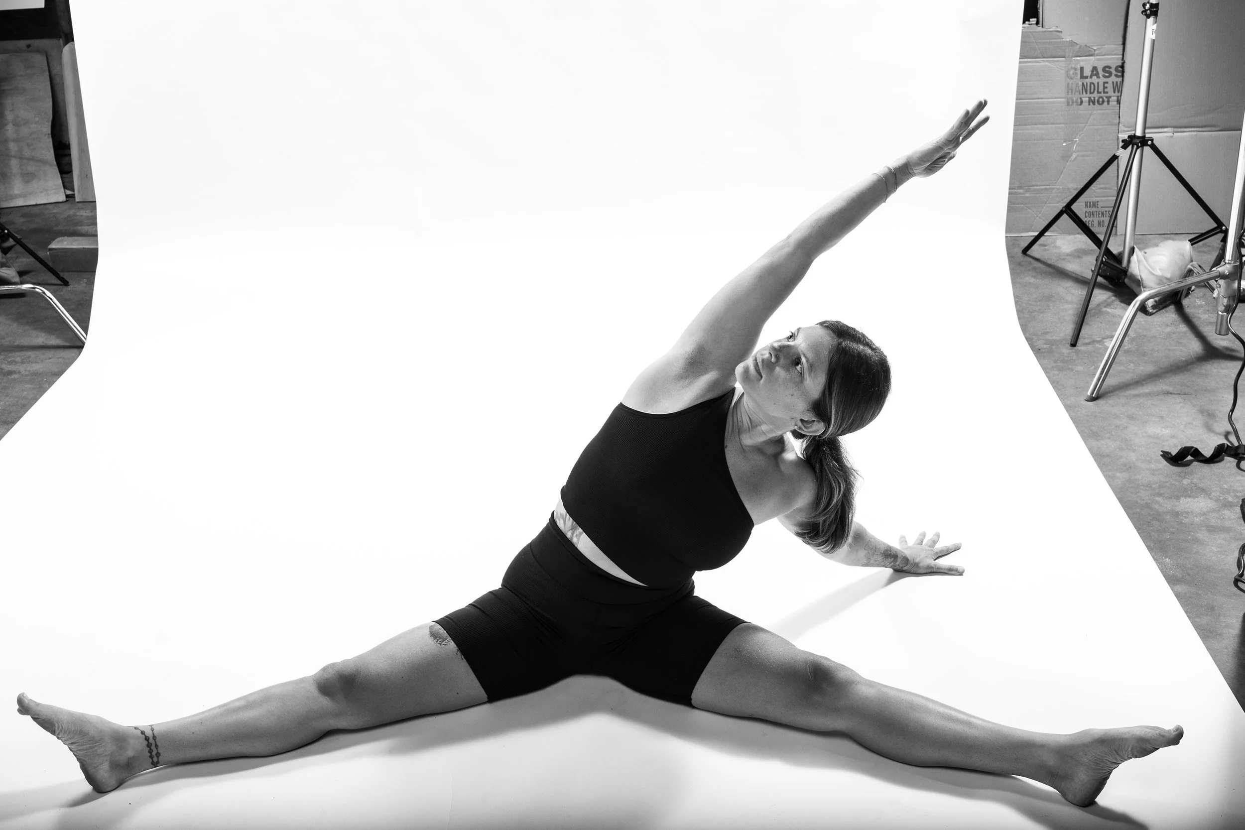 A woman doing a seated side stretch yoga pose on a white backdrop in a studio.
