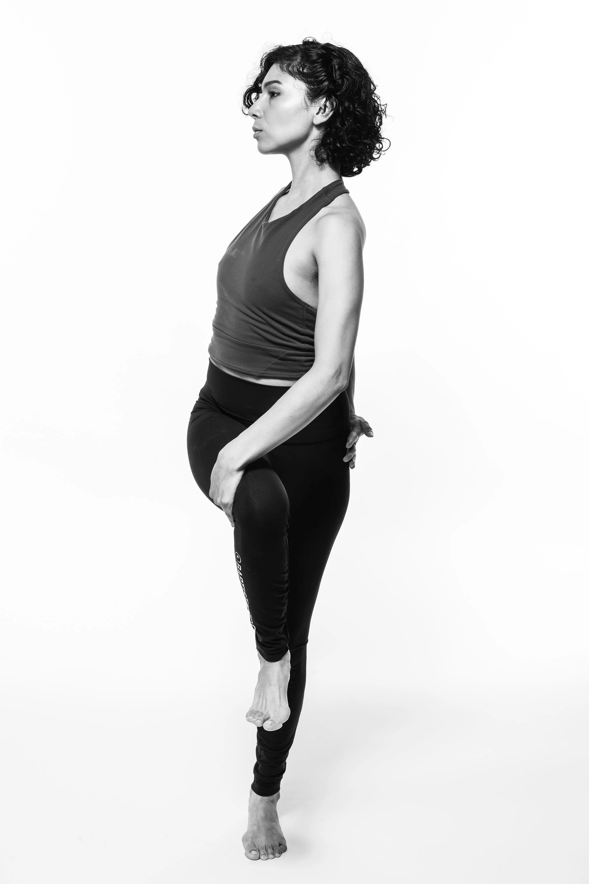 A woman practicing yoga, standing on one leg with her hands behind her back, in a studio with a plain background.