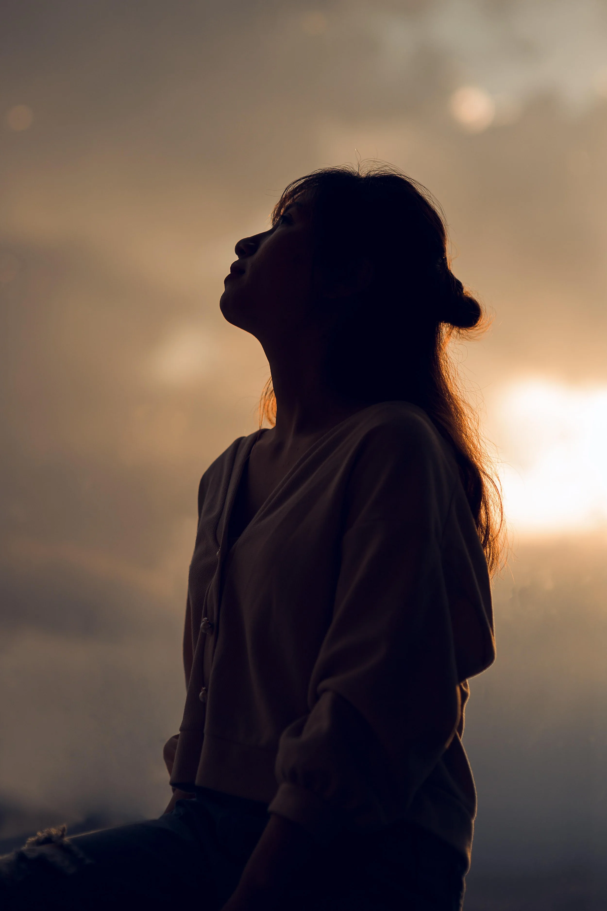 Schatten einer Frau bei Sonnenuntergang, sitzend mit Blick nach oben, im Hintergrund bewölkter Himmel mit warmem Licht.