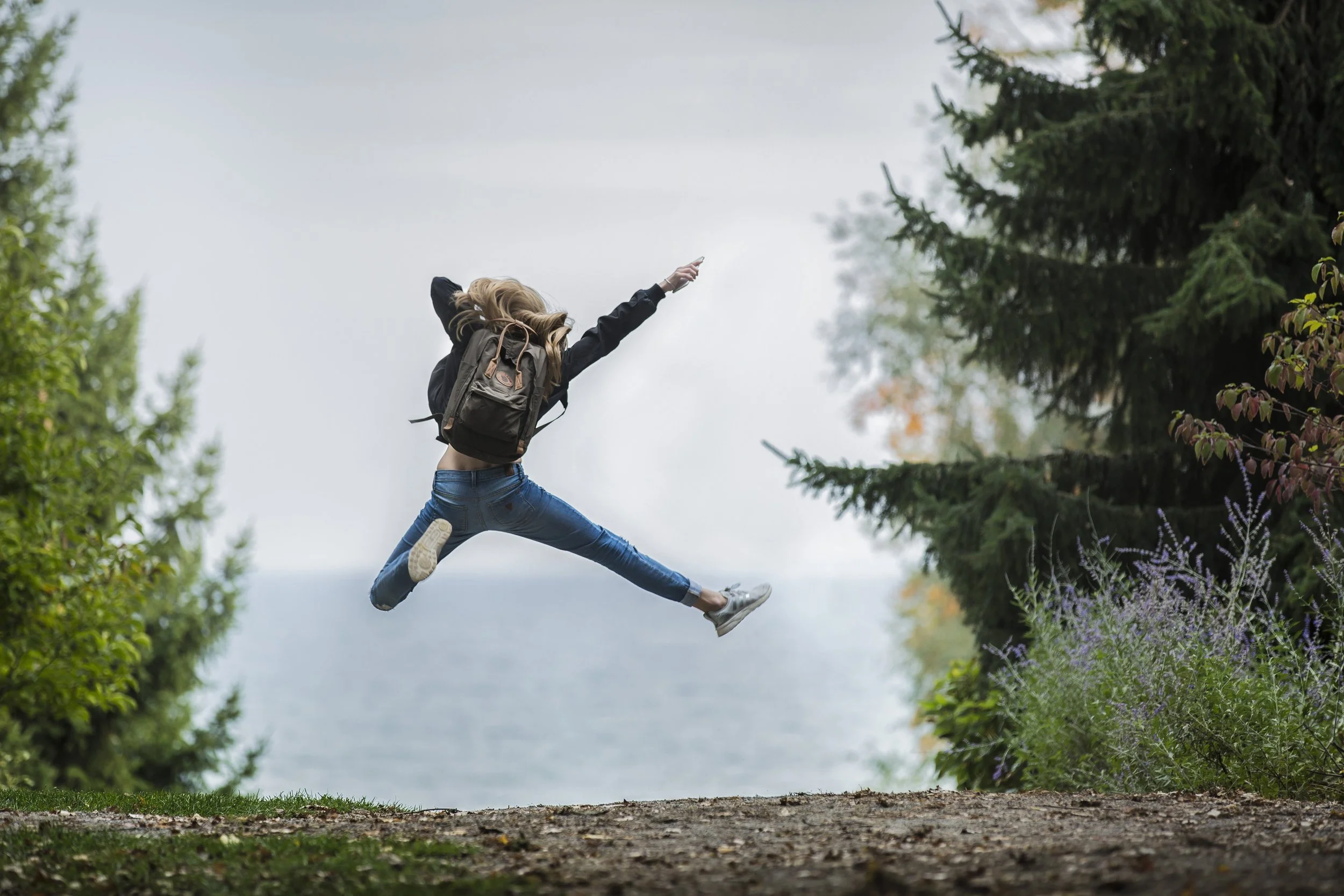 Junge Frau springt in der Natur zwischen Bäumen und Büschen in die Luft, trägt Rucksack und sportliche Kleidung.