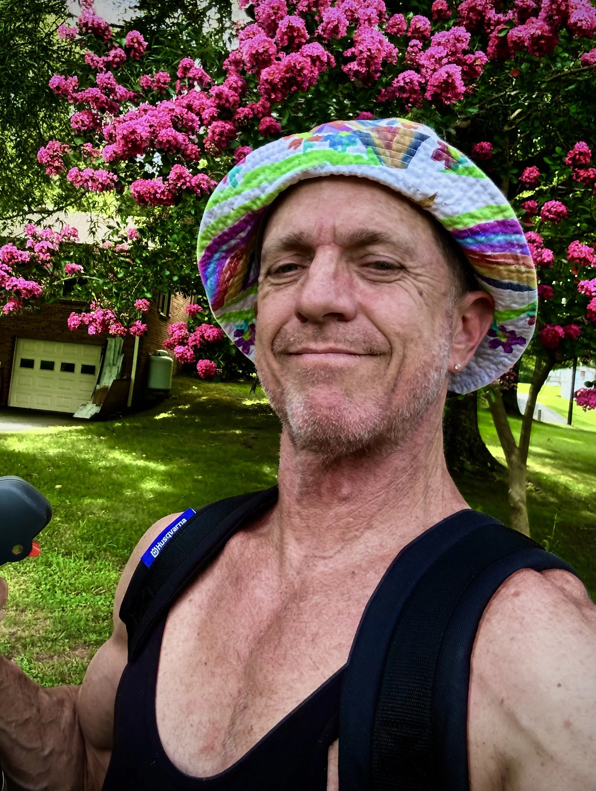A smiling man wearing a colorful bucket hat and a black tank top takes a selfie outdoors in front of a pink flowering tree.
