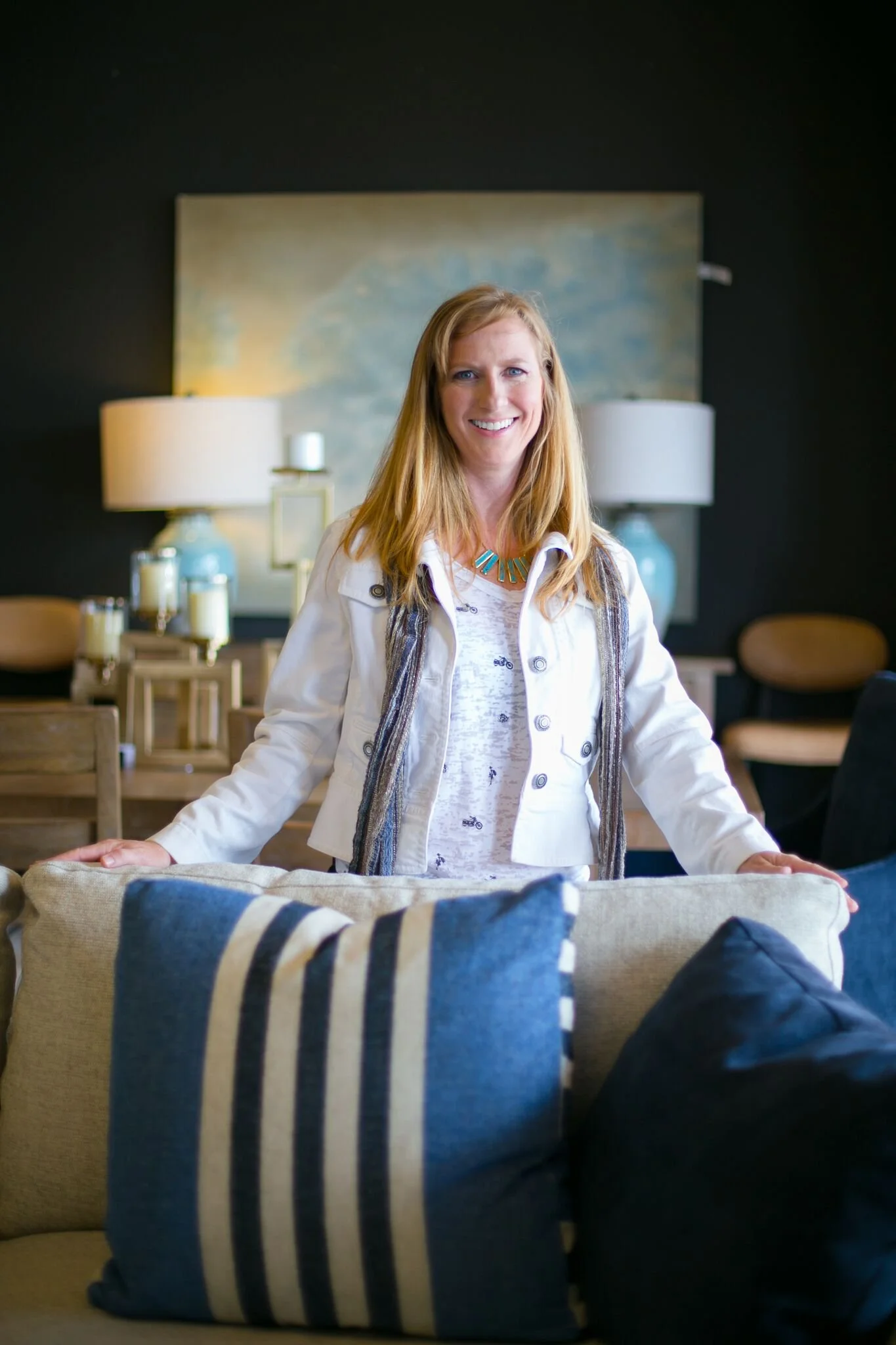 A woman with long red hair smiling and standing behind a beige sofa, decorated with blue and dark pillows, in a living room with dark walls, lamps, and table decor in the background.