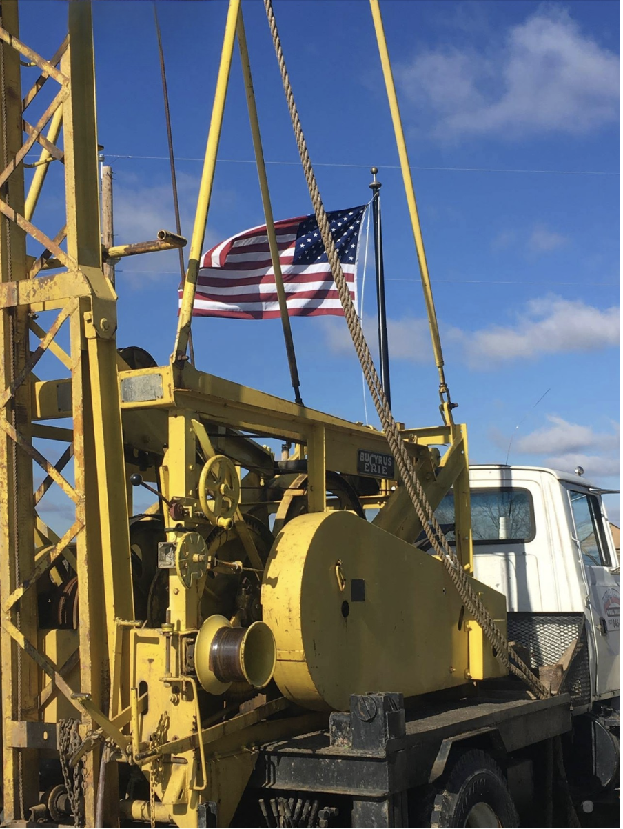 A yellow drilling rig truck with an American flag flying against a blue sky with clouds.