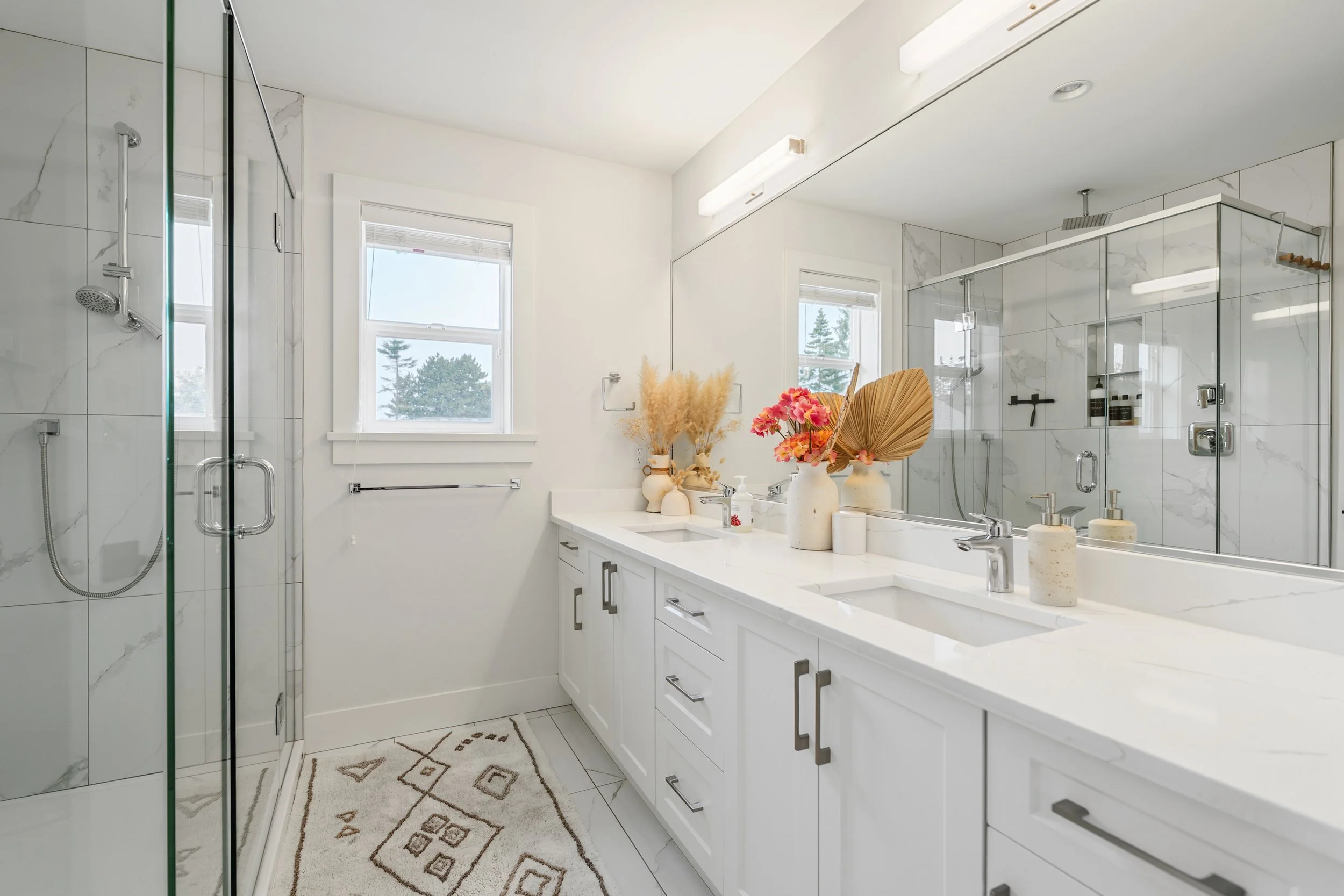 Bright modern primary bathroom with double white vanity, large mirror, and glass walk-in tile shower professionally photographed for real estate listing.