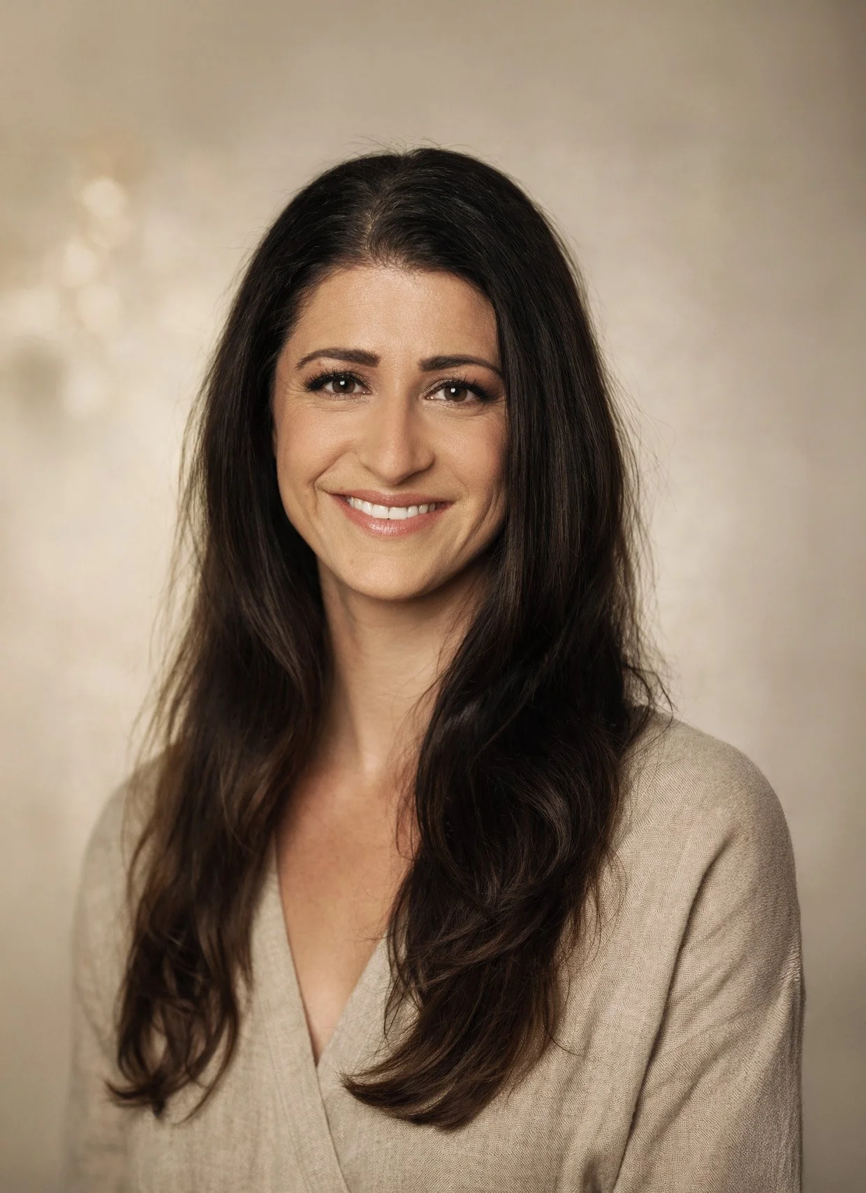 Eine Frau mit dunklem, langem, gewelltem Haar, leicht lächelnd, trägt einen beigen Pullover, im Hintergrund eine neutrale, leicht strukturierte Wand.