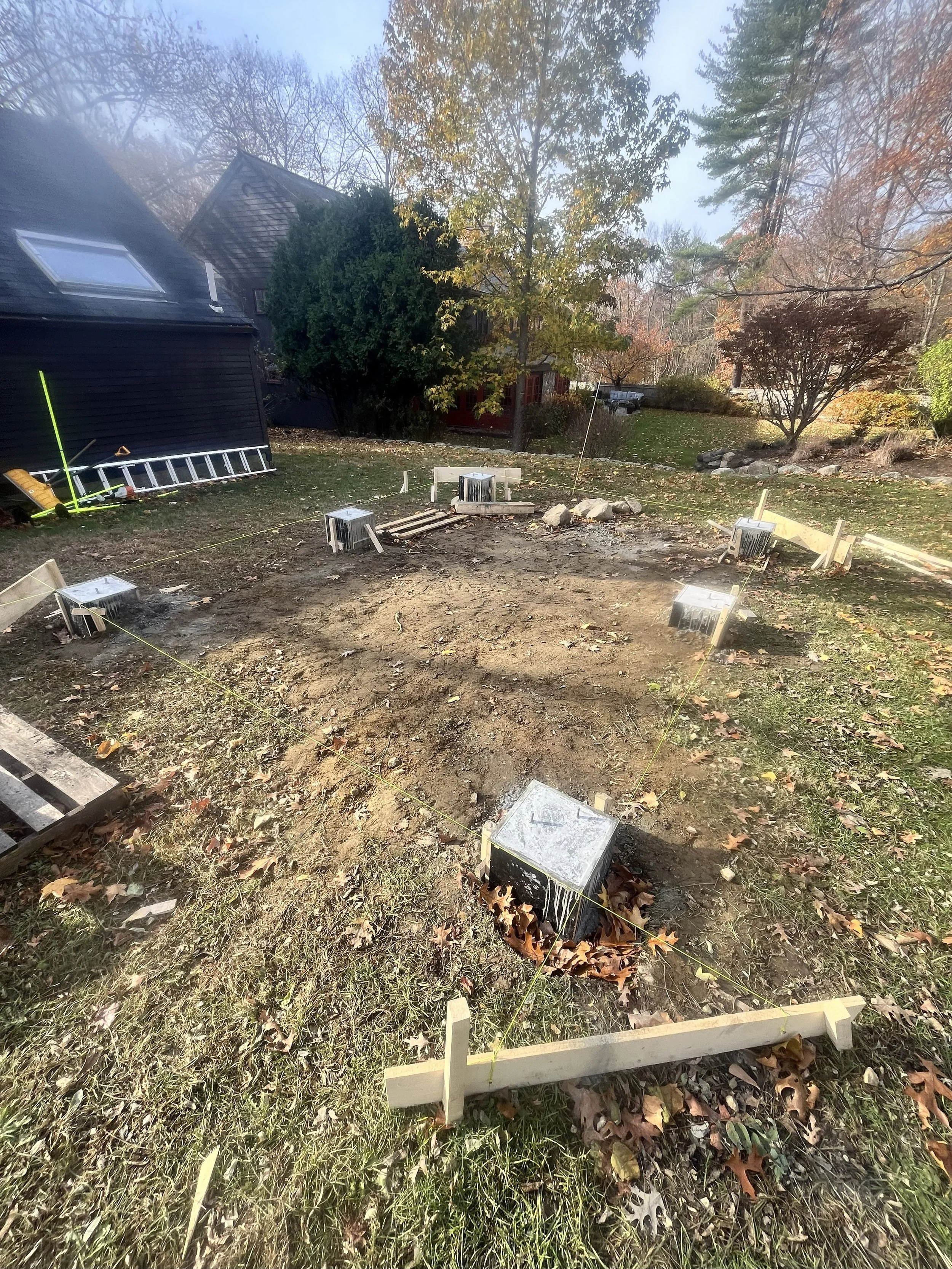 Home construction site with exposed foundation, wooden stakes, and leveling lines in a backyard, surrounded by trees with fall foliage.