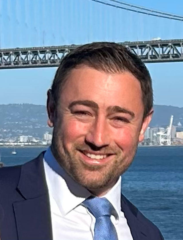 A man in a suit smiling outdoors with a bridge and water in the background.