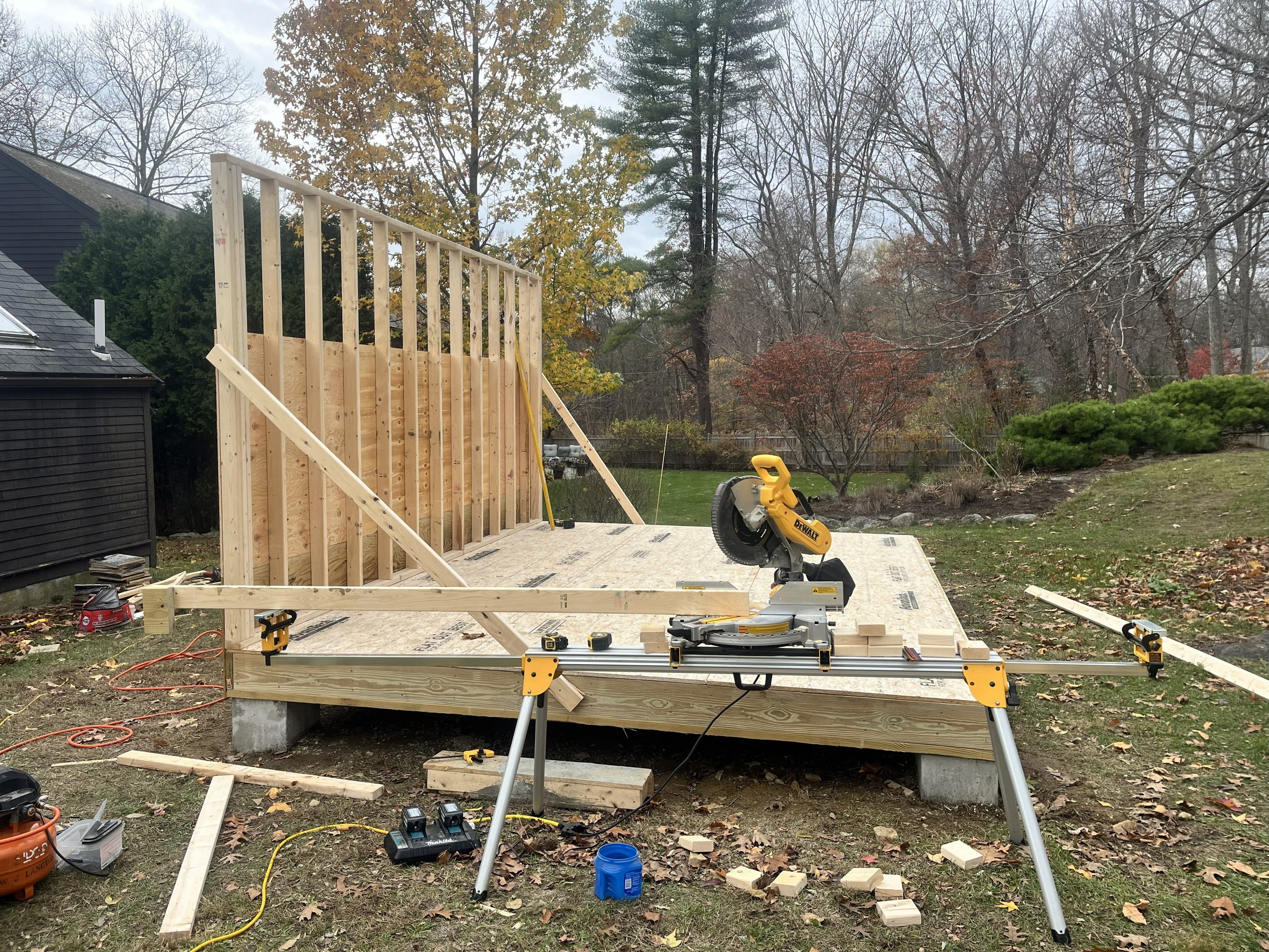 Construction site with a wooden deck in progress, including a mounted miter saw, with trees and a house in the background.