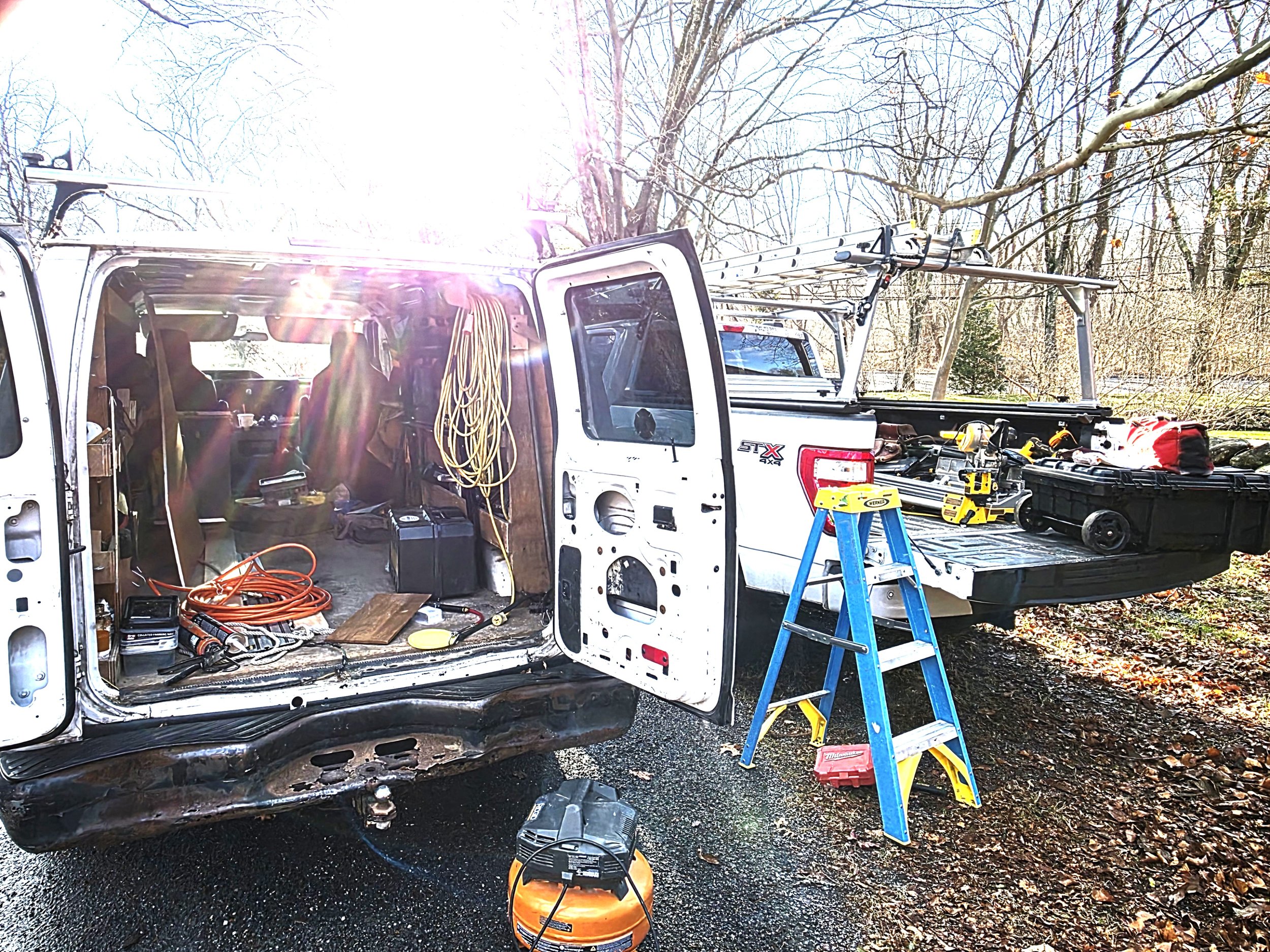 An open work van with tools, extension cords, and equipment inside. The back of the van has ladders, a toolbox, and power tools. A step ladder is set up outside, and the scene is outdoors with trees and sunlight.