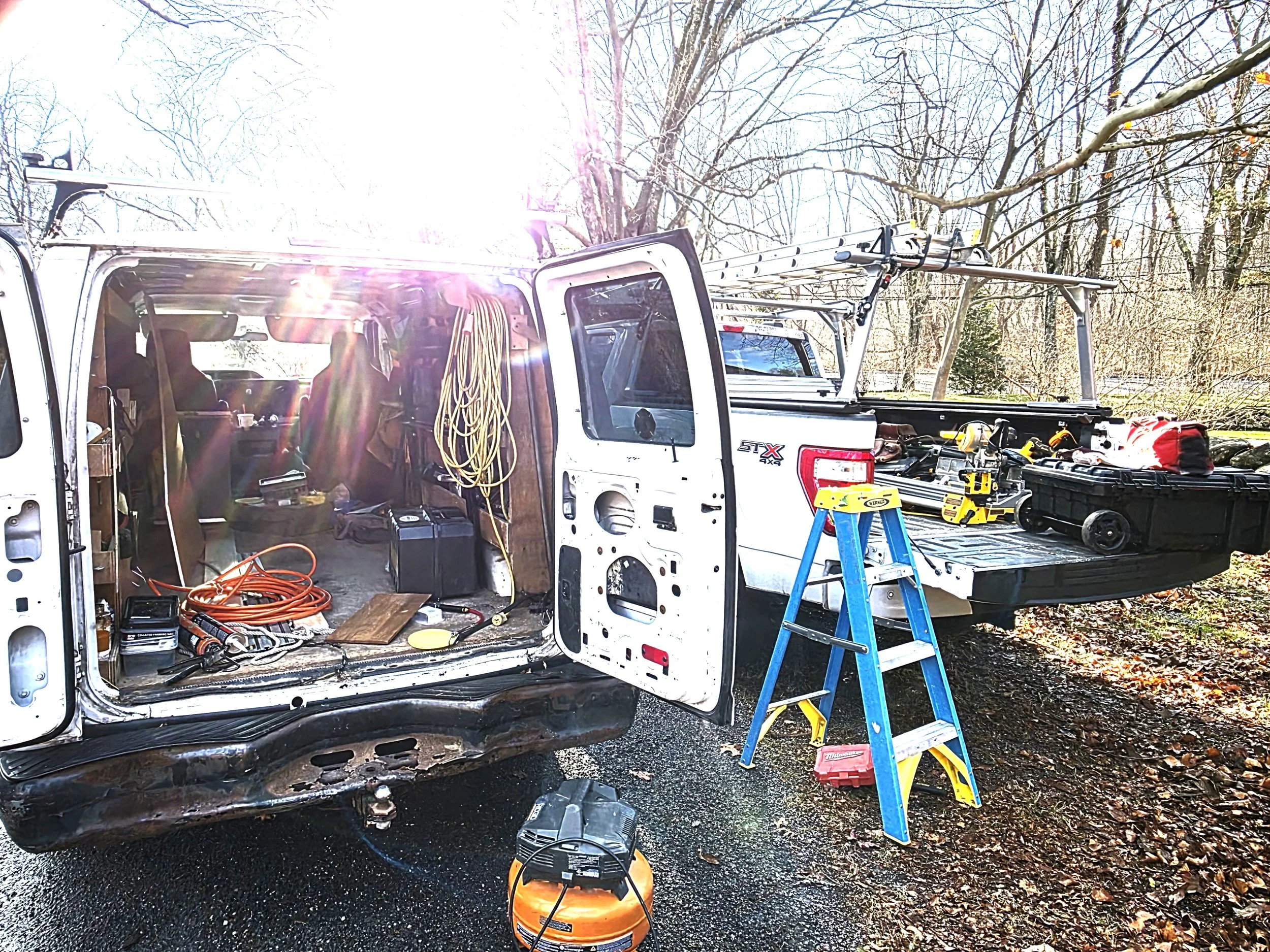 An open work van with tools, extension cords, and equipment inside. The back of the van has ladders, a toolbox, and power tools. A step ladder is set up outside, and the scene is outdoors with trees and sunlight.