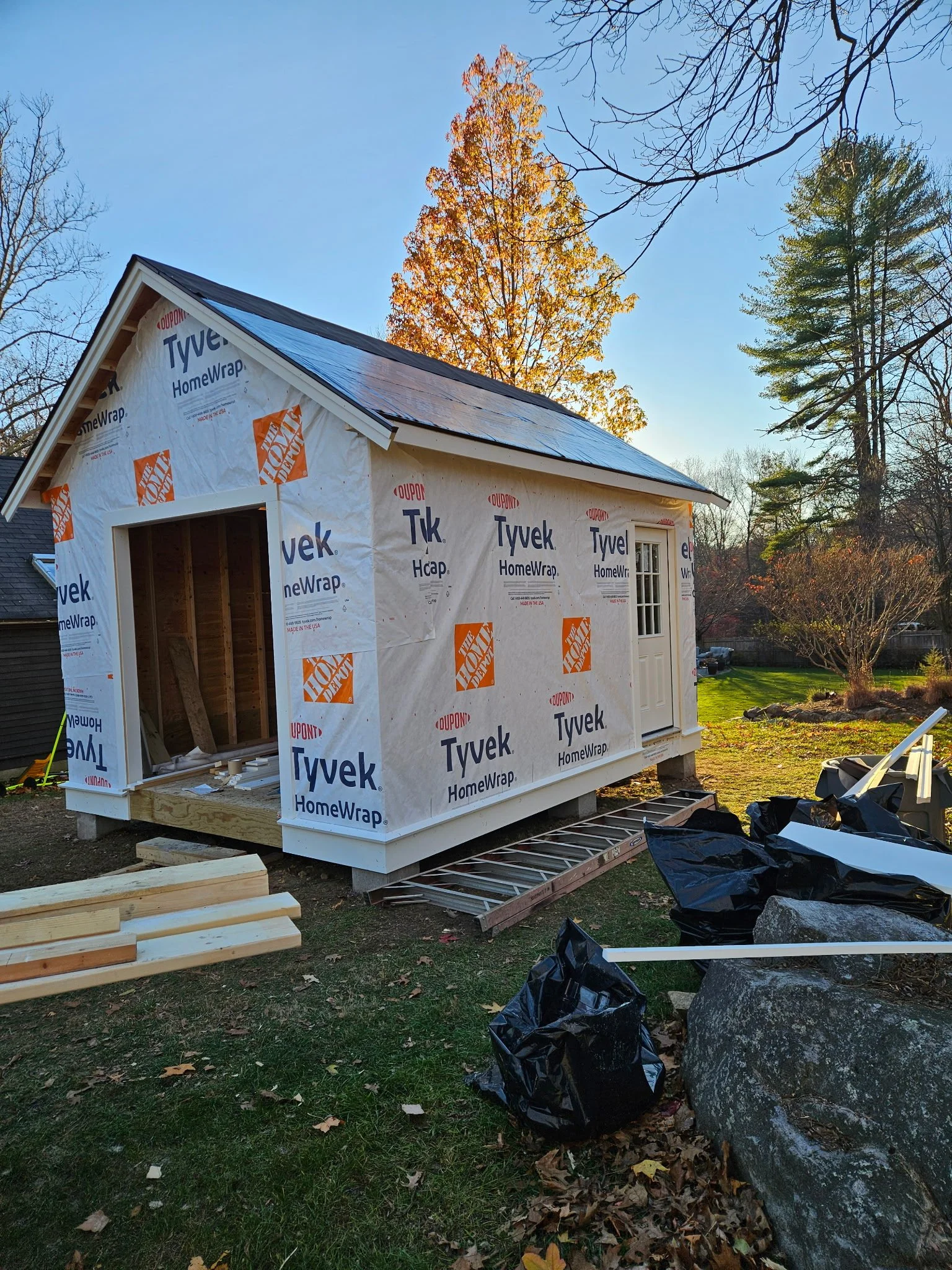 Backyard of a house under construction with partial exterior wall wrapping, ladder, black trash bags, and construction materials on the grass, surrounded by trees with autumn foliage.