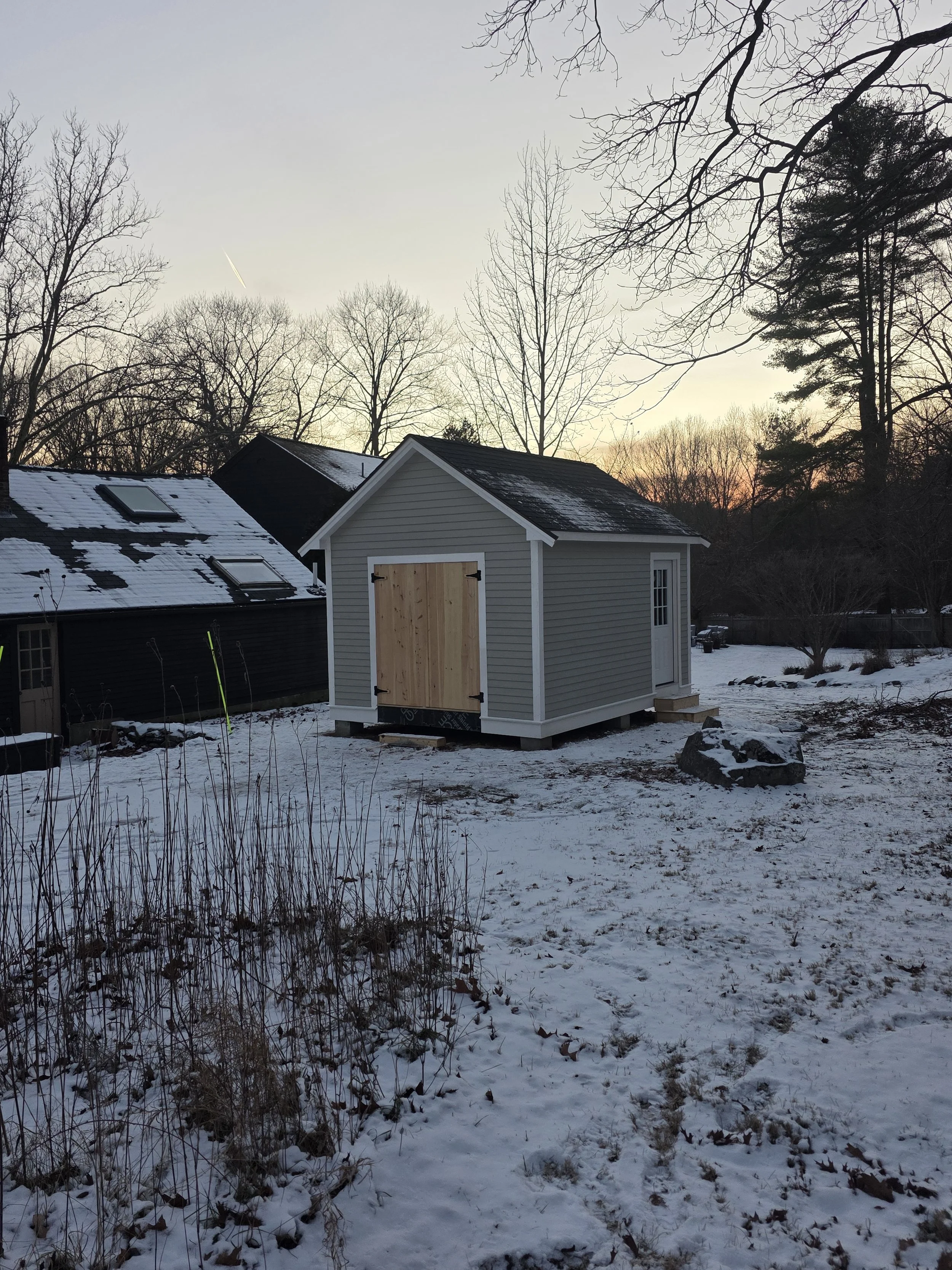 A small gray shed with white trim and a wooden door in a snowy backyard, with trees and neighboring houses in the background at sunset.