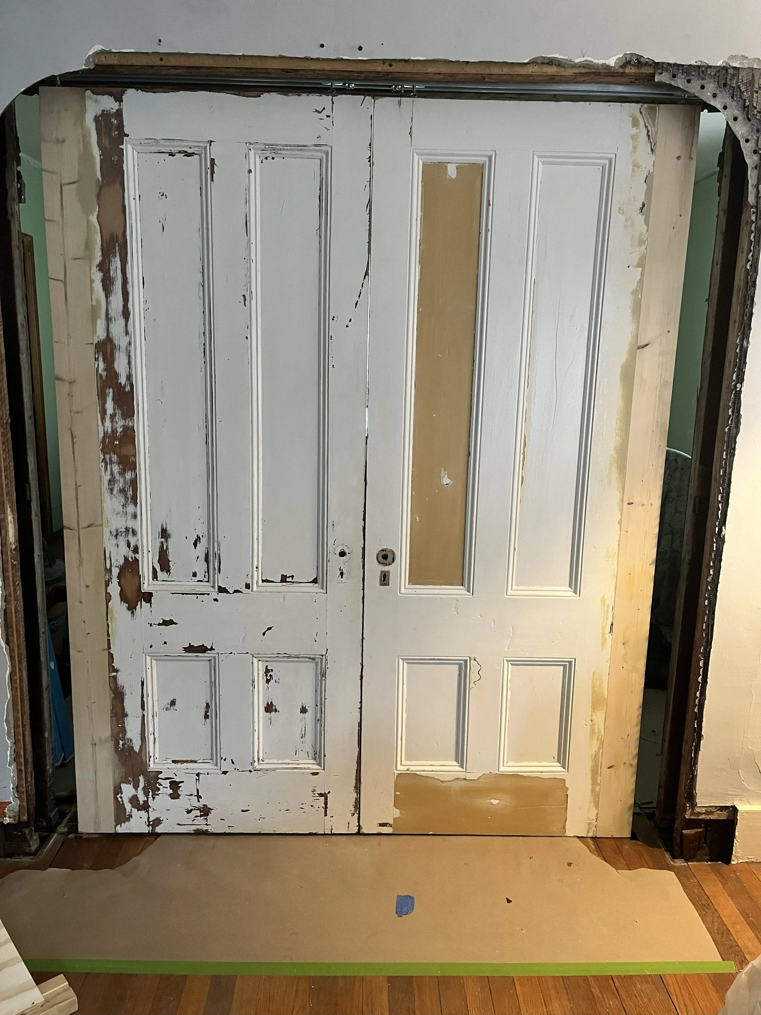 Old double wooden doors with peeling white paint, partially restored, with some panels sanded or primed, and patches of exposed wood or primer, leaning against a wall in a room under renovation.