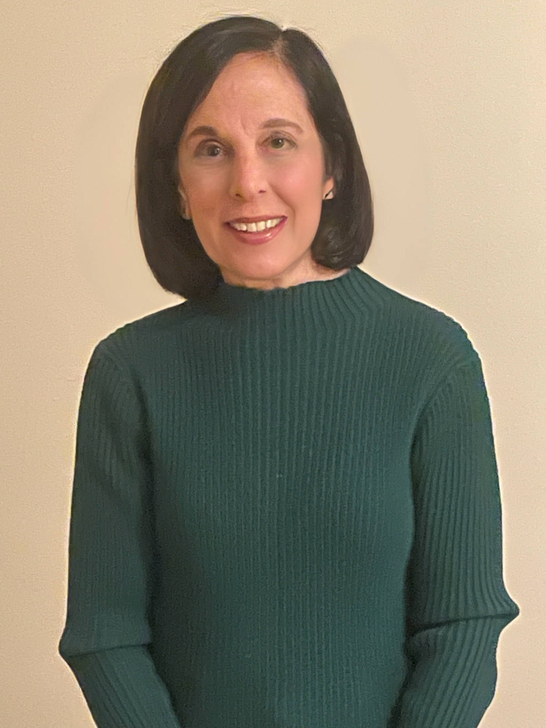 A woman with shoulder-length dark hair, smiling, wearing a green ribbed sweater, standing against a plain light-colored wall.