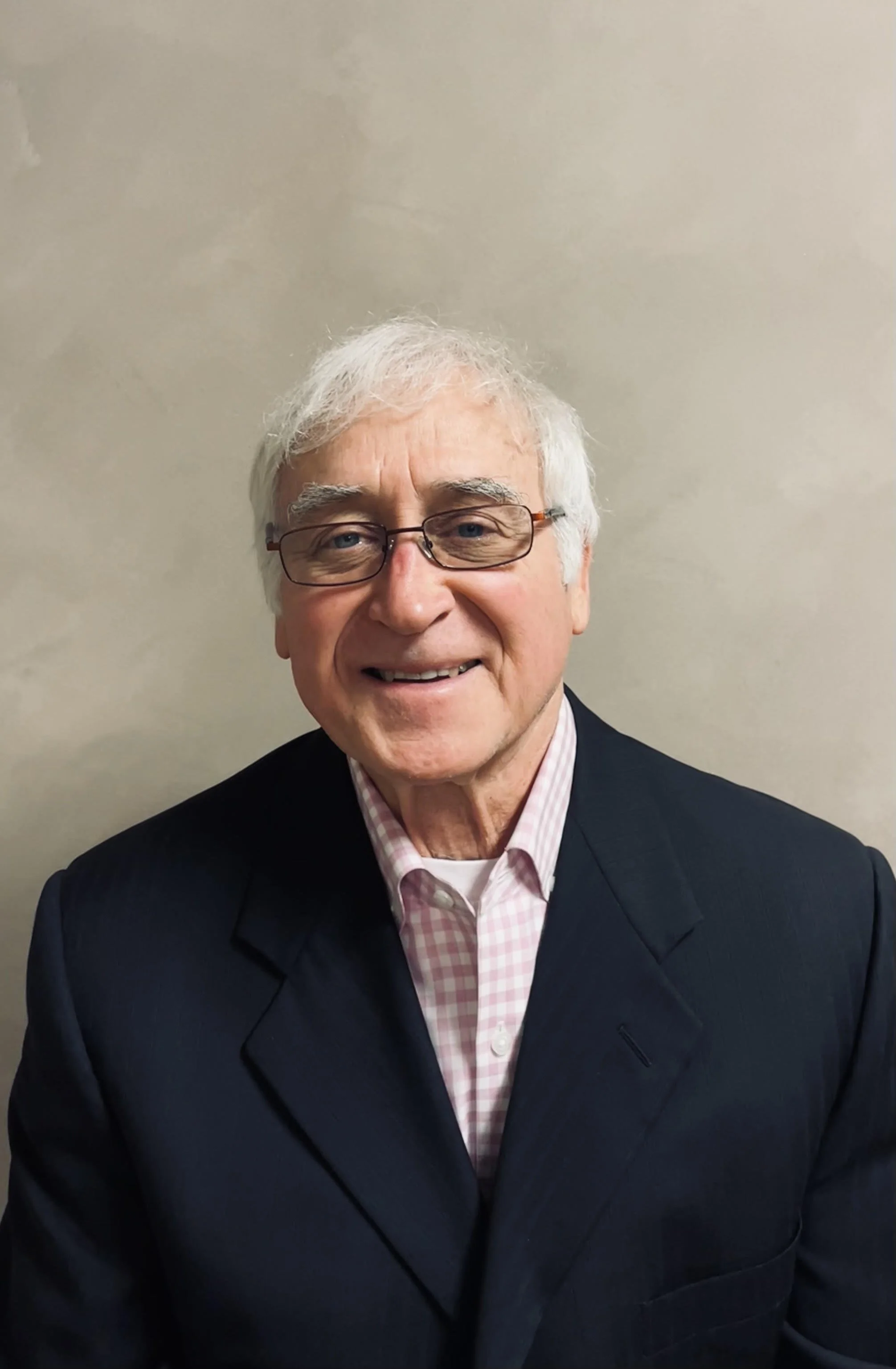 A smiling older man with white hair, glasses, wearing a dark suit jacket and a pink checkered shirt, standing against a plain light-colored wall.