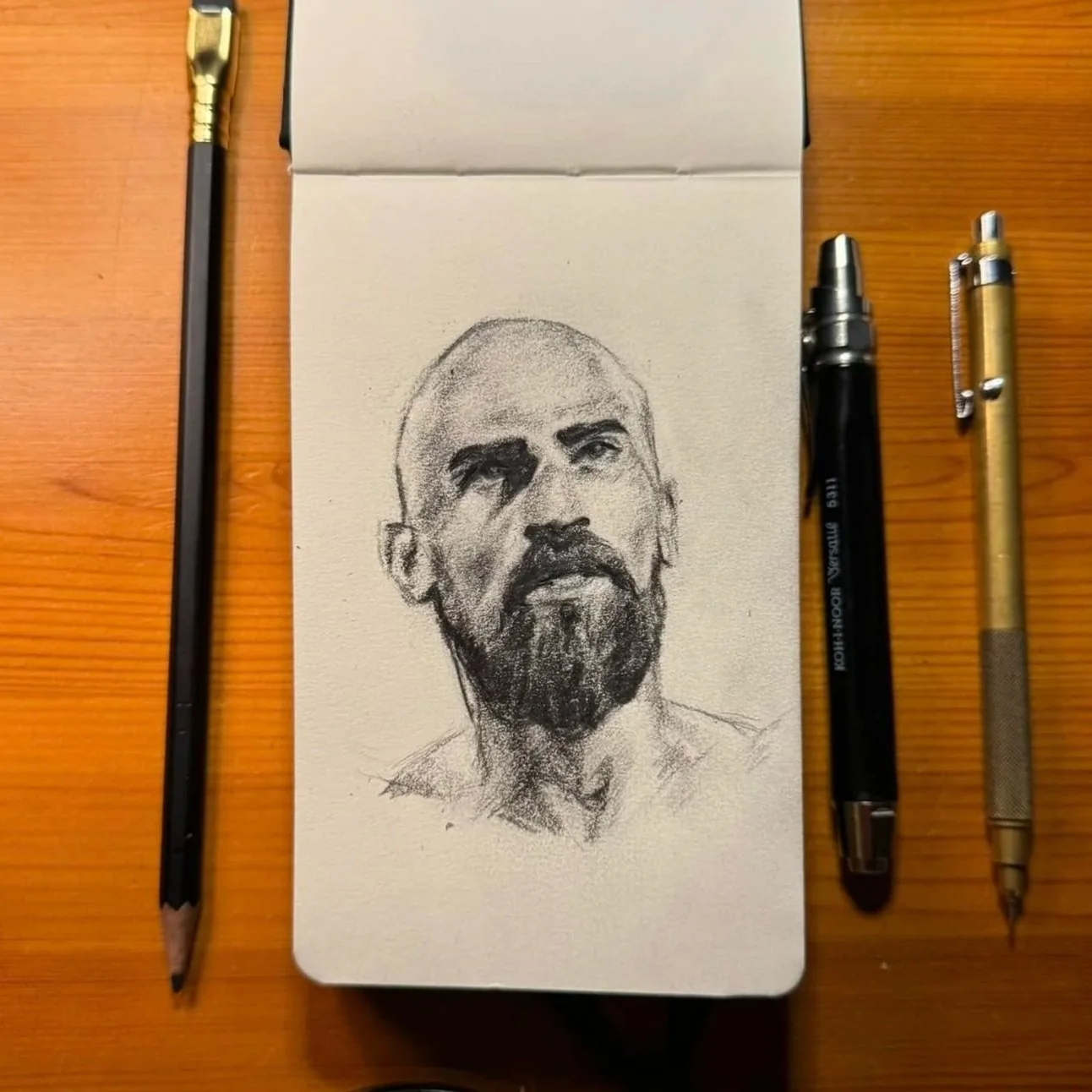 Sketch of a man with a serious expression, short hair, a beard, and a mustache on a small sketchbook, surrounded by three pens and a pencil, on a wooden surface.