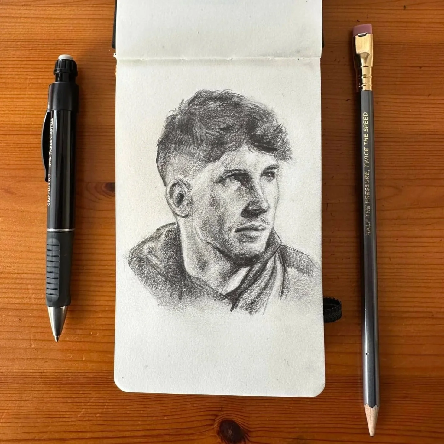 A detailed pencil sketch of a young man with short, wavy hair, wearing a jacket, on a white sketchbook page. Two pens are placed on either side of the sketch, on a wooden surface.