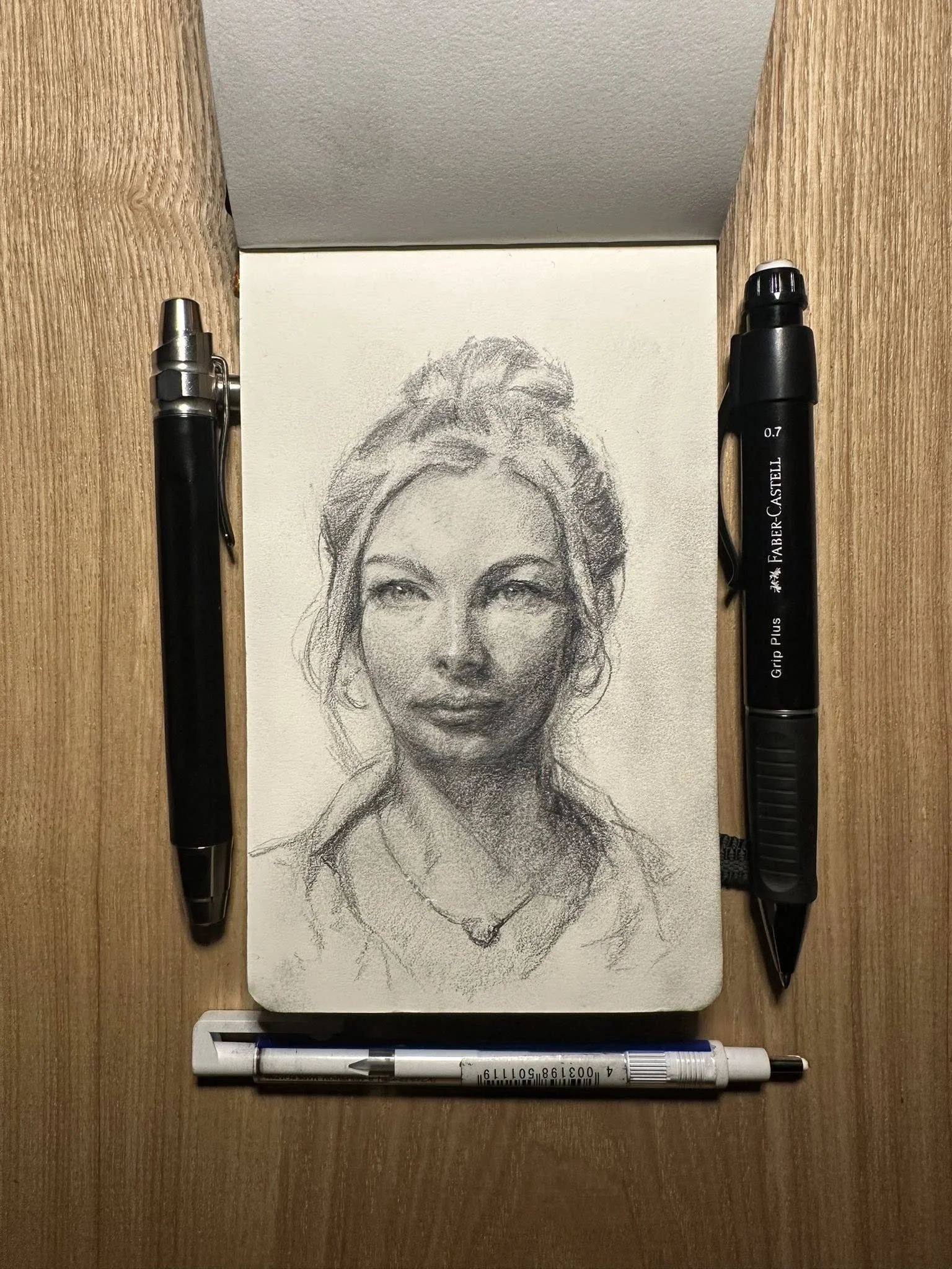 Sketch of a woman with light-colored hair styled in a bun, wearing a necklace, on white paper, flanked by two black pens and a white pen with black details on a wooden surface.