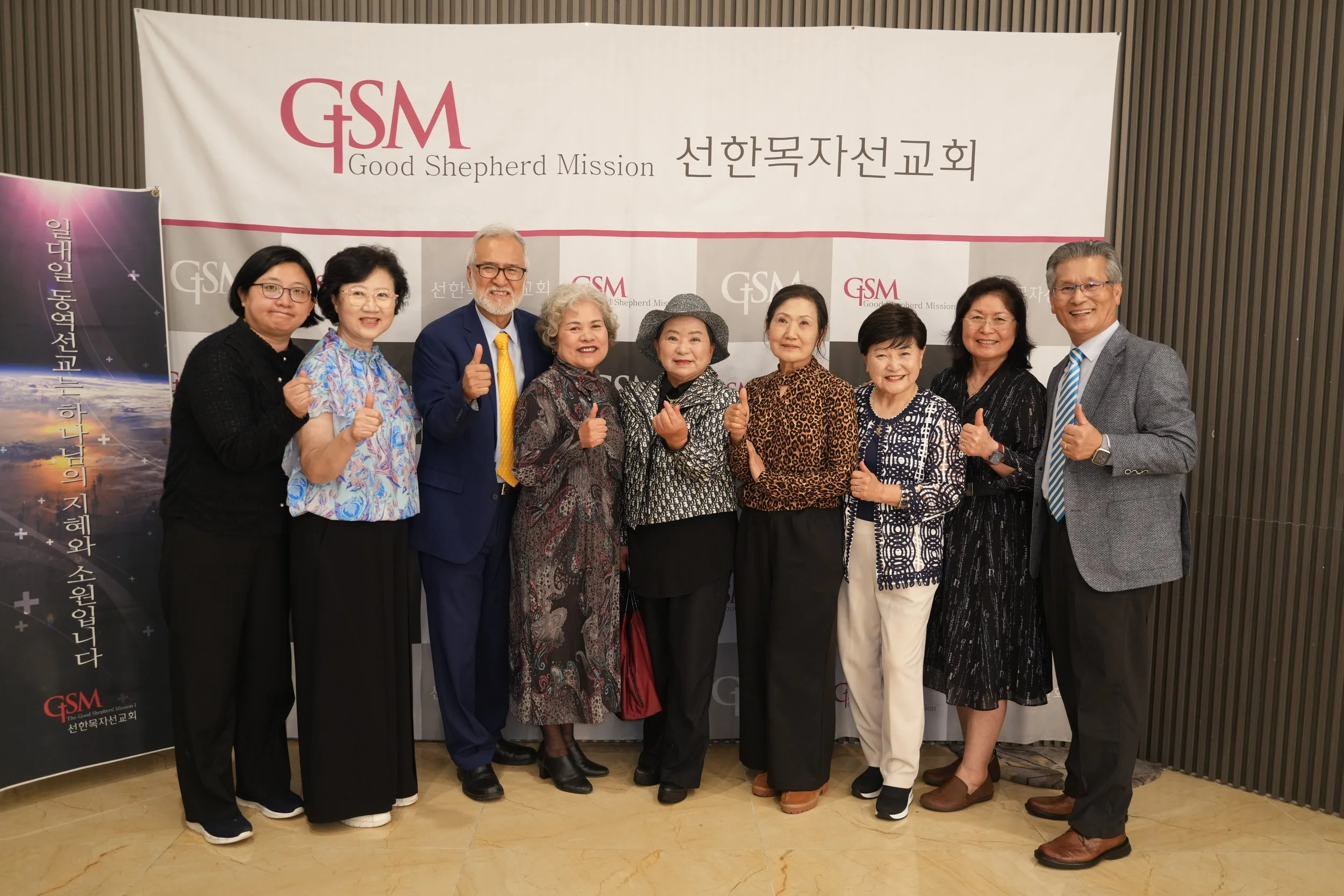 Group of ten diverse adults standing in front of a large banner with 'G+S M Good Shepherd Mission' written on it, at an indoor event. They are smiling, some making thumbs-up gestures, and are dressed in business casual and formal attire.