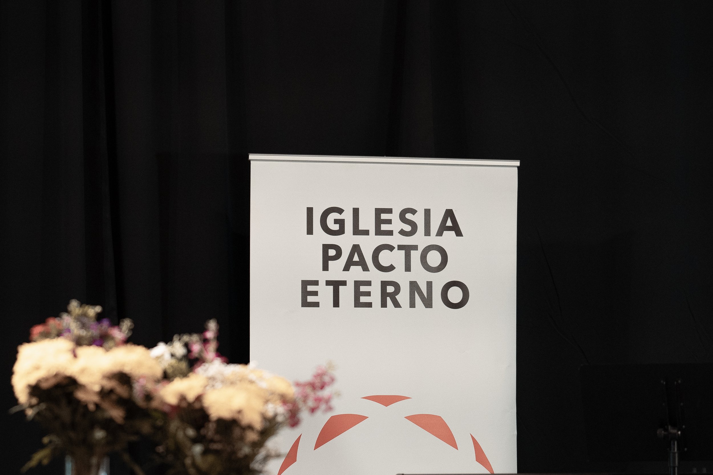 Púlpito con cartel que dice 'Iglesia Pacto Eterno' y un arreglo floral al frente, fondo negro.