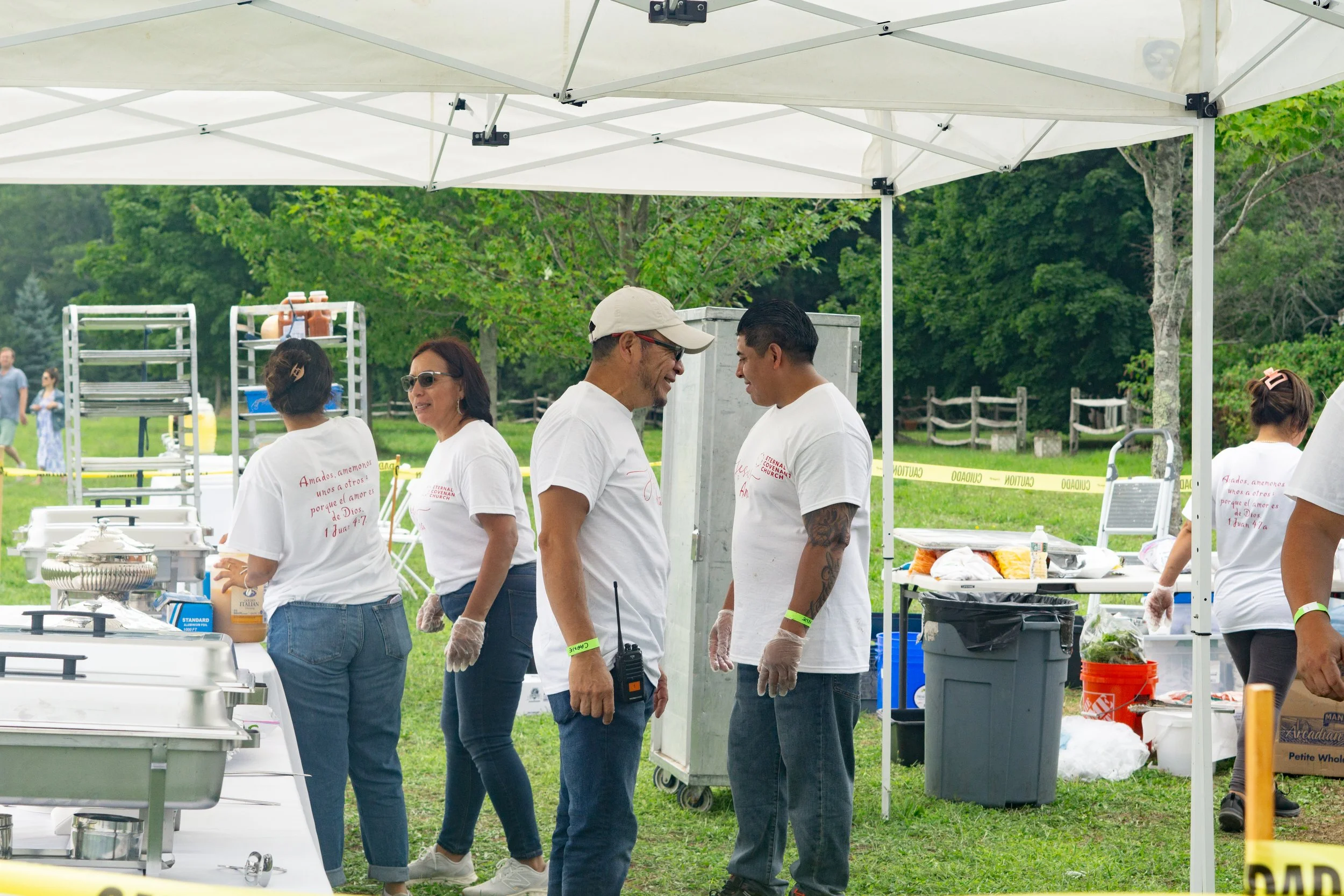 Personas trabajando en la preparación de comida en un evento al aire libre con carpa y áreas de grass. Algunas personas usan guantes y camisetas blancas con texto en rojo.