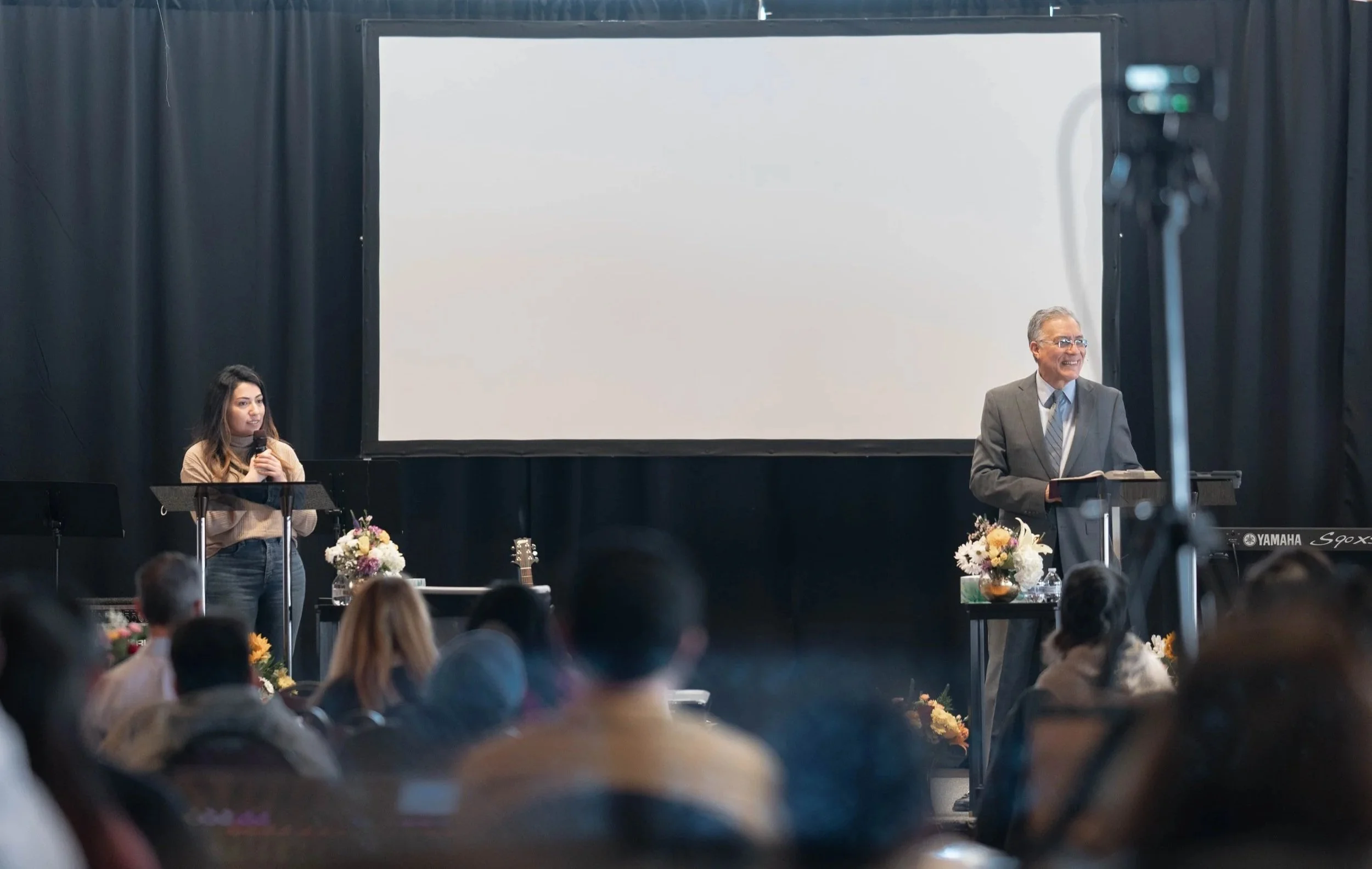 Una mujer y un hombre en un escenario durante una presentación con público. La mujer sostiene un micrófono y está a la izquierda, mientras que el hombre, vestido con traje, está a la derecha tras un atril. Hay decoraciones florales en las mesas cerca del escenario, y en el fondo hay una pantalla grande en blanco.