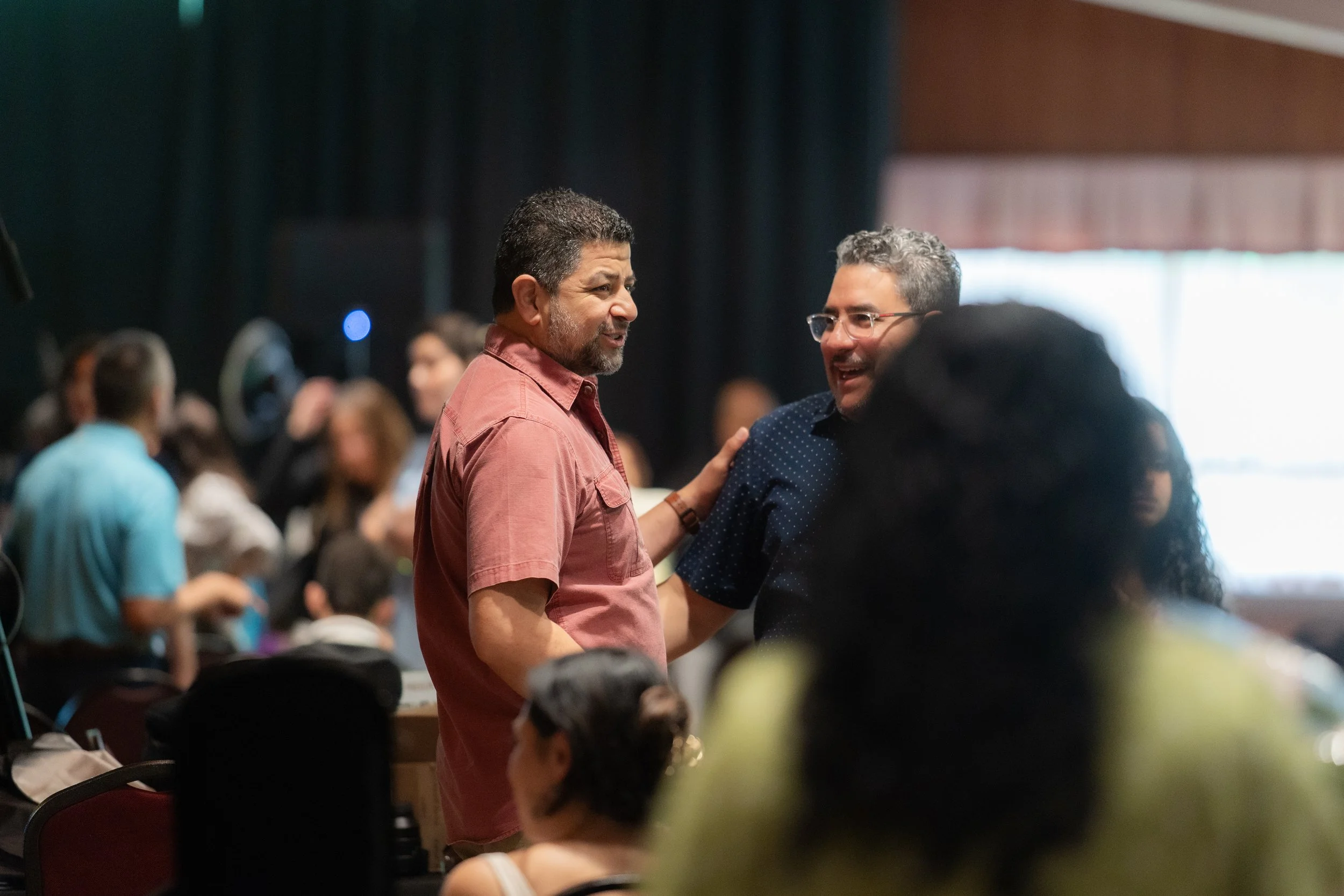 Dos hombres conversando en un evento, uno con camiseta rosa y el otro con camiseta azul con puntos, en un ambiente concurrido con varias personas en el fondo.