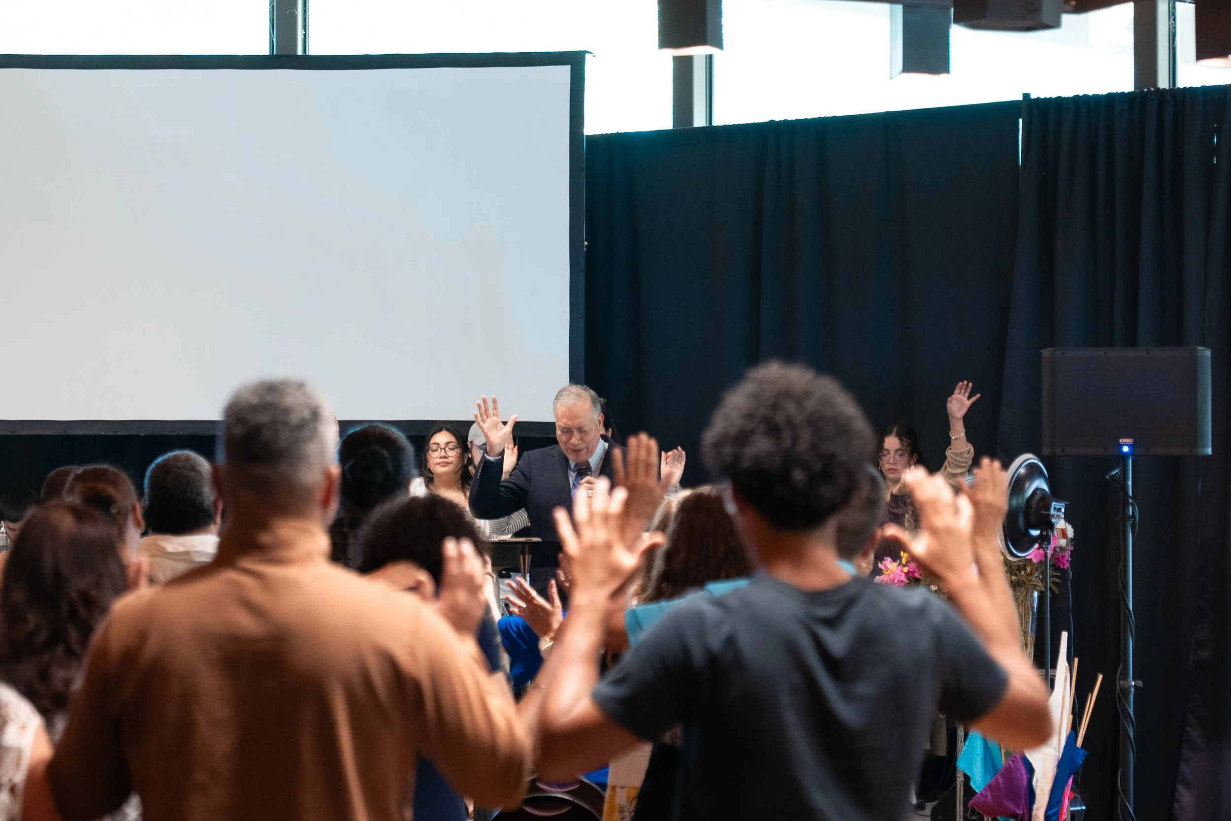 Personas en una audiencia con muchas manos levantadas en un evento o conferencia, con un orador en el fondo frente a una pantalla grande.