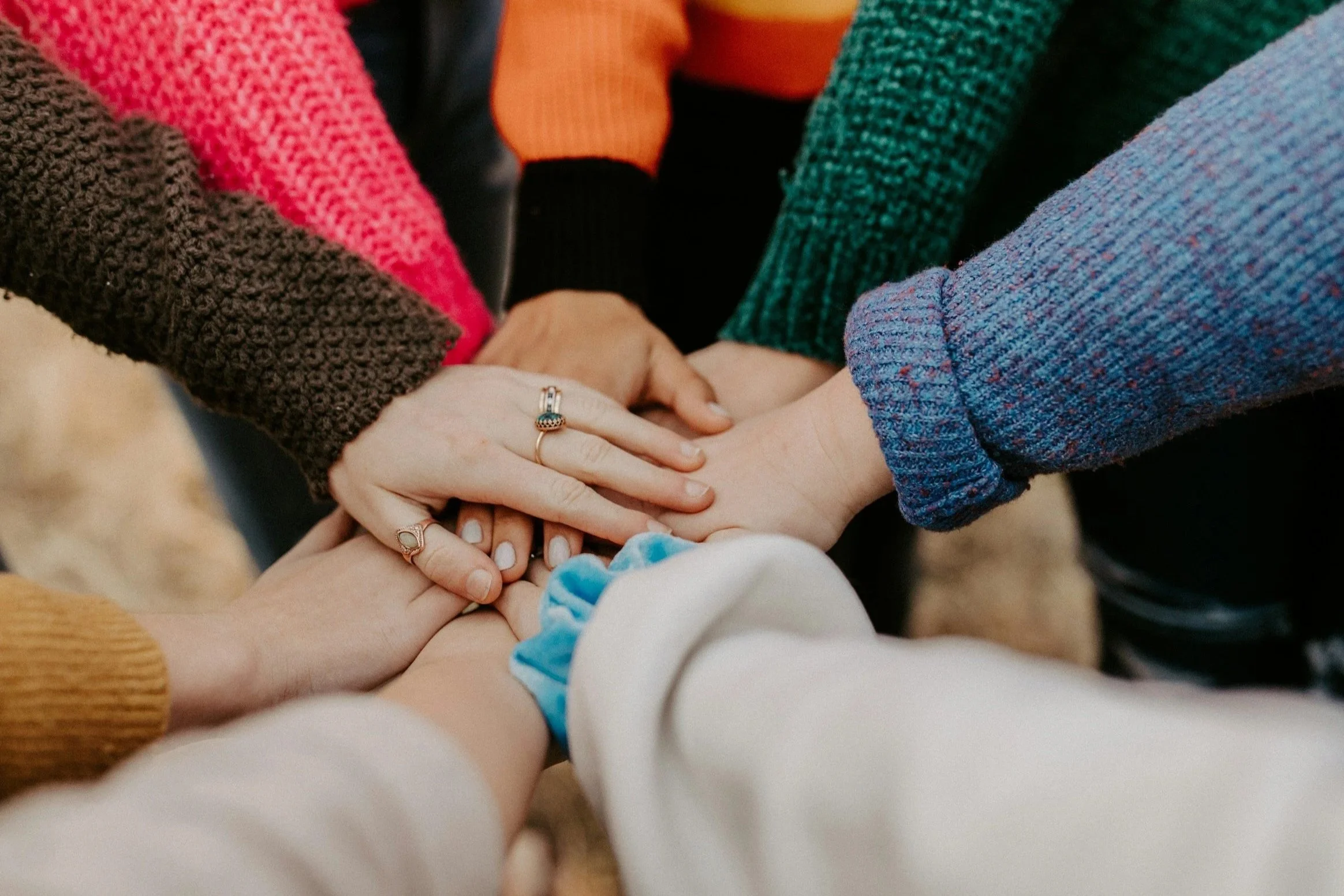 A group of women stacking their hands together, showing diversity with different skin tones and wearing colorful sweaters.