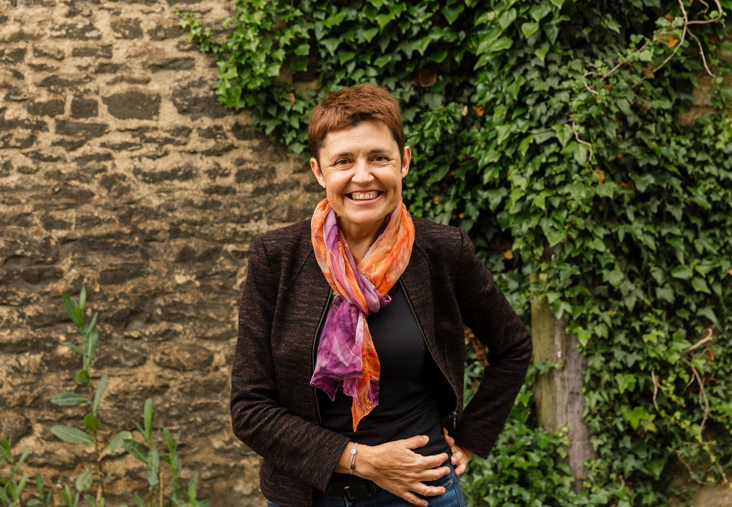Rosa  Arias-Yague, a woman with short brown hair wearing a dark jacket, colorful scarf, and black top, standing outdoors in front of a stone wall with green ivy while smiling.