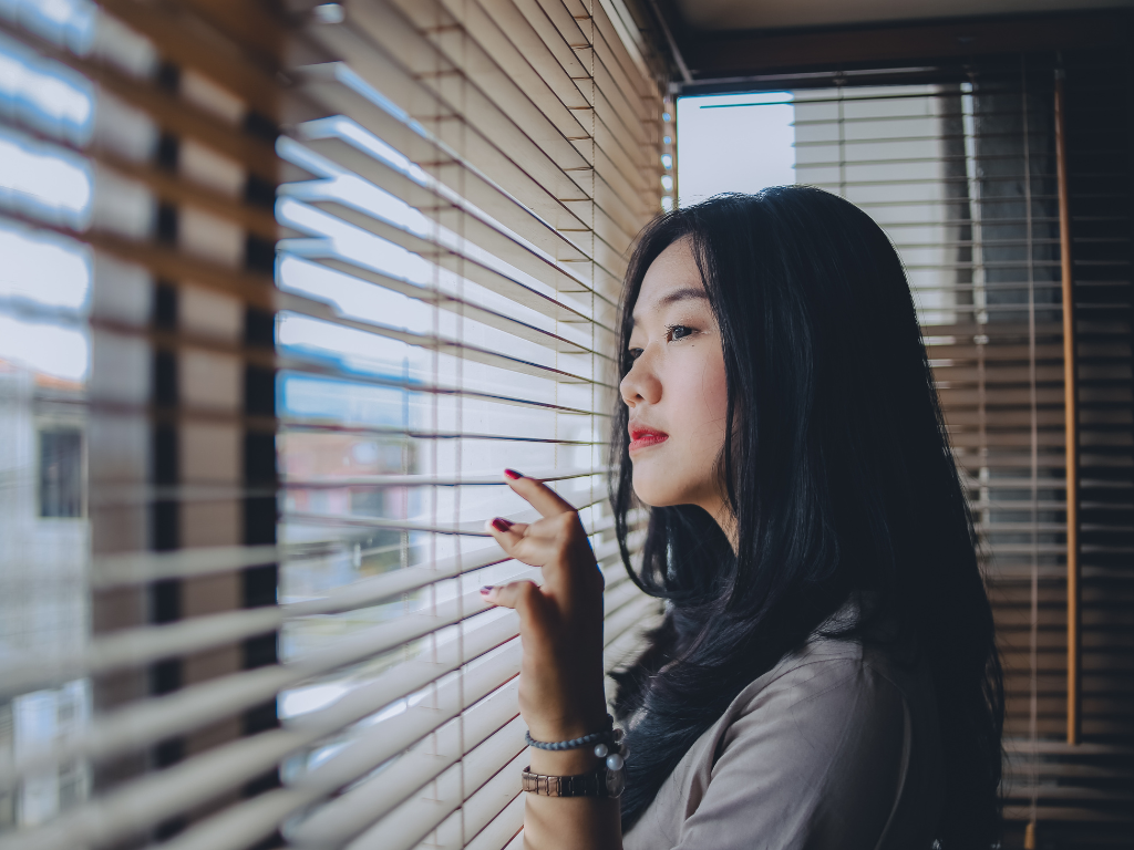 An Asian woman with black hair looking out through window blinds.