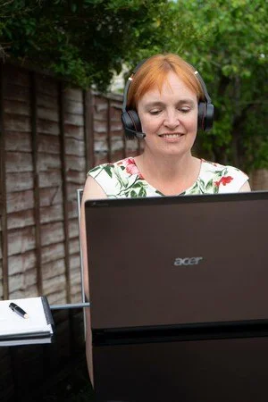 A woman with short red hair wearing headphones, smiling, sitting outdoors in front of a laptop, with a wooden fence and green trees in the background.