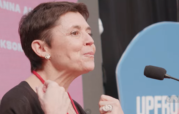 Rosa Arias-Yague (short brown hair and silver earrings) standing while speaking at a conference.