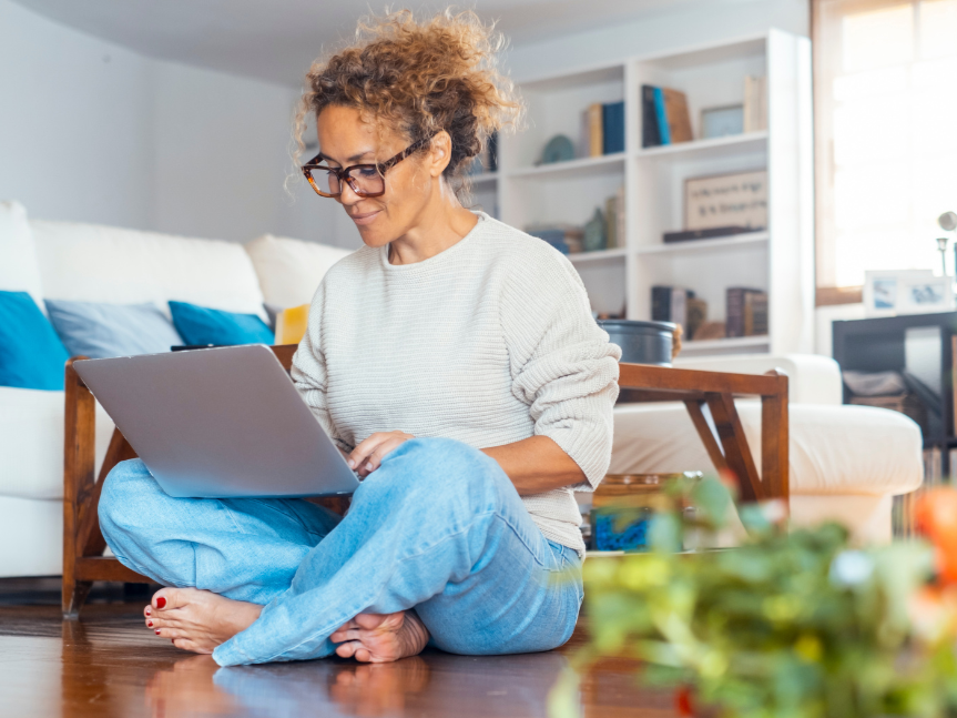 A Black woman with curly hair and glasses sitting cross-legged on the floor at home, using a laptop.