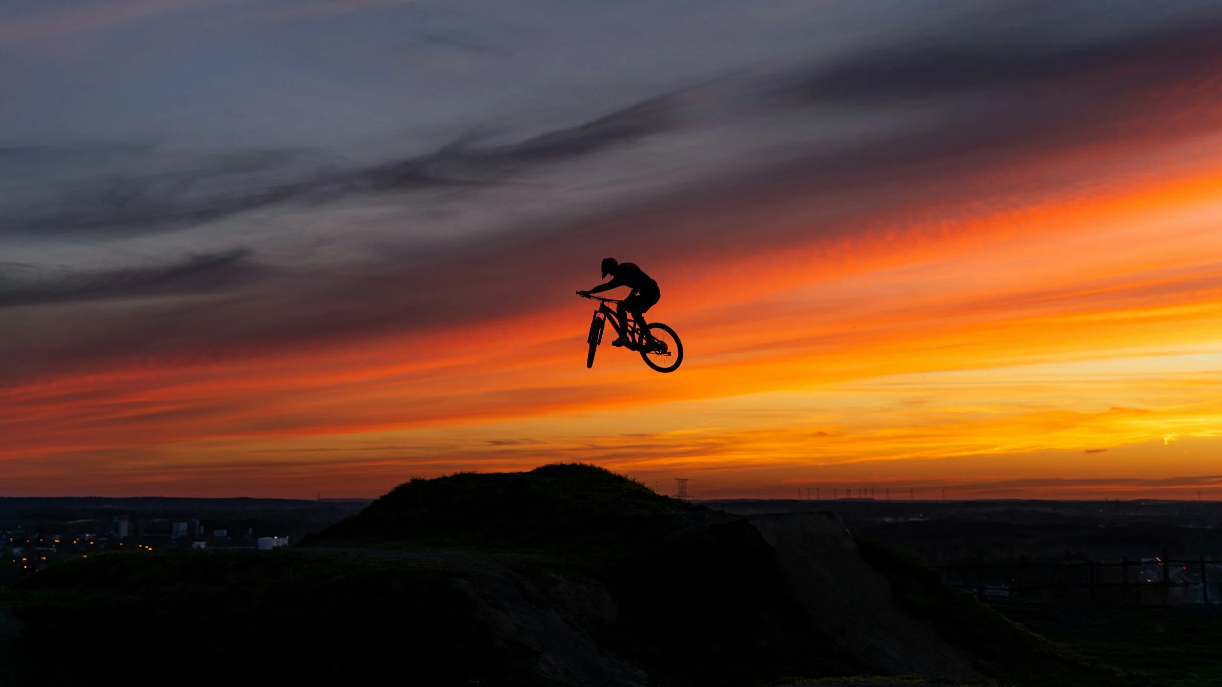 A silhouette of a person doing a mid-air bicycle stunt at sunset, with a hill and a colorful sky in the background.