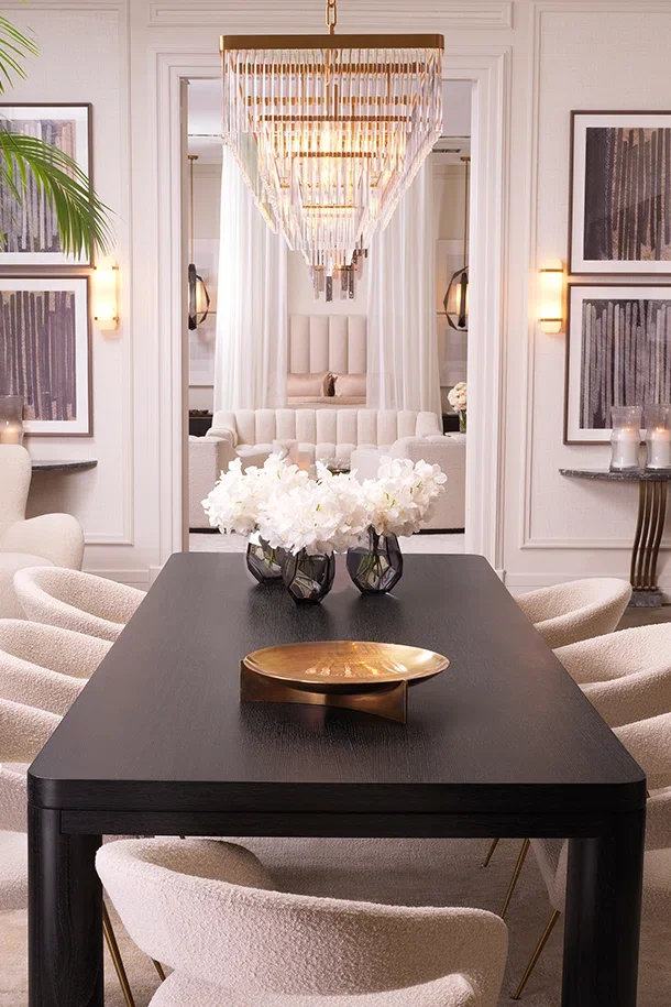 A dining room with a black table, beige upholstered chairs, a modern gold decorative bowl, and a centerpiece of white flowers in black vases. There is a large, elegant chandelier hanging above the table, with white walls adorned with framed artwork. In the background, a cozy sitting area features a white tufted sofa, soft lighting, and white curtains.