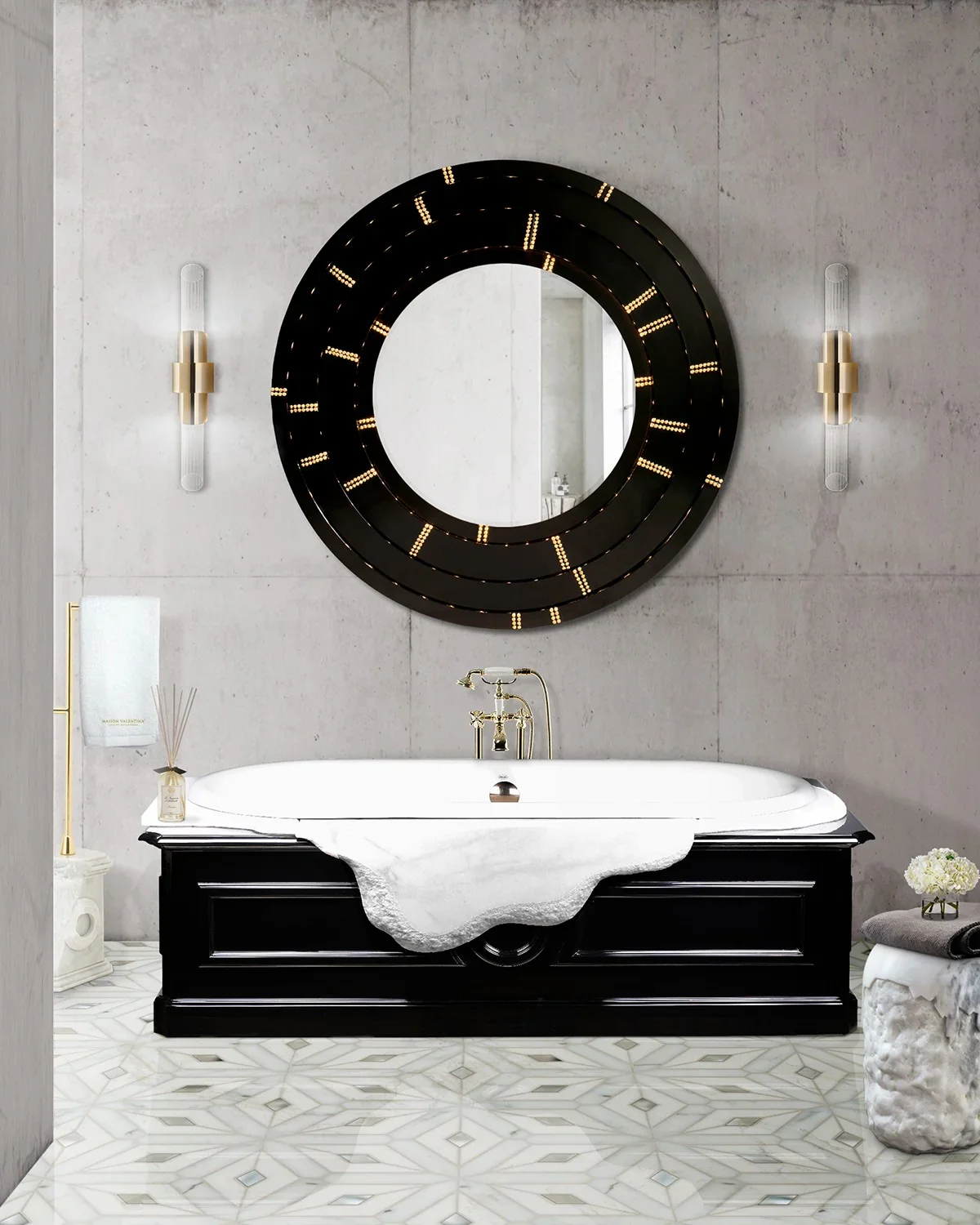 A contemporary bathroom featuring a black-framed bathtub with white marble accent, gold fixtures, a large round mirror with lighting, and gray stone walls.
