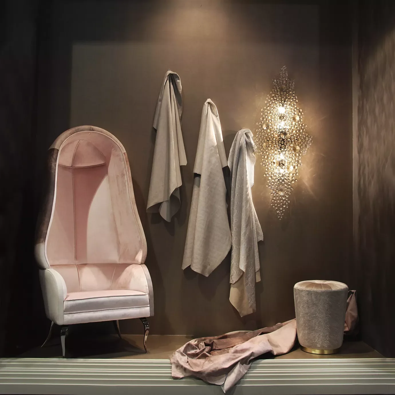 A luxurious pink velvet high-back armchair with silver legs, four beige hanging coats, a gray round ottoman with a gold base, a draped pink fabric on the floor, and a decorative wall sconce made of numerous small circular mirrors emitting warm light.