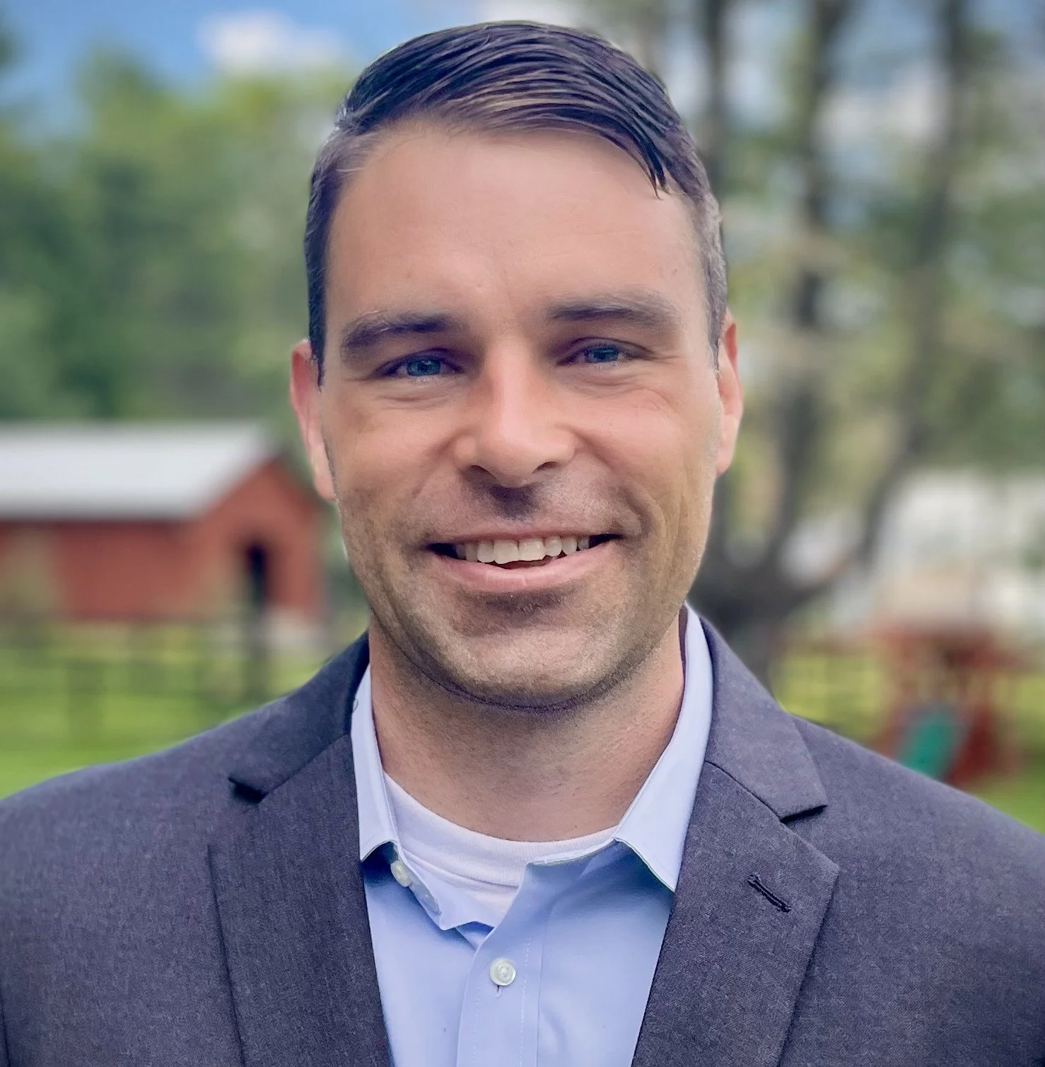 A man with dark hair, blue eyes, and light skin in a gray blazer and light blue shirt, smiling outdoors with a blurred background of trees and a red building.