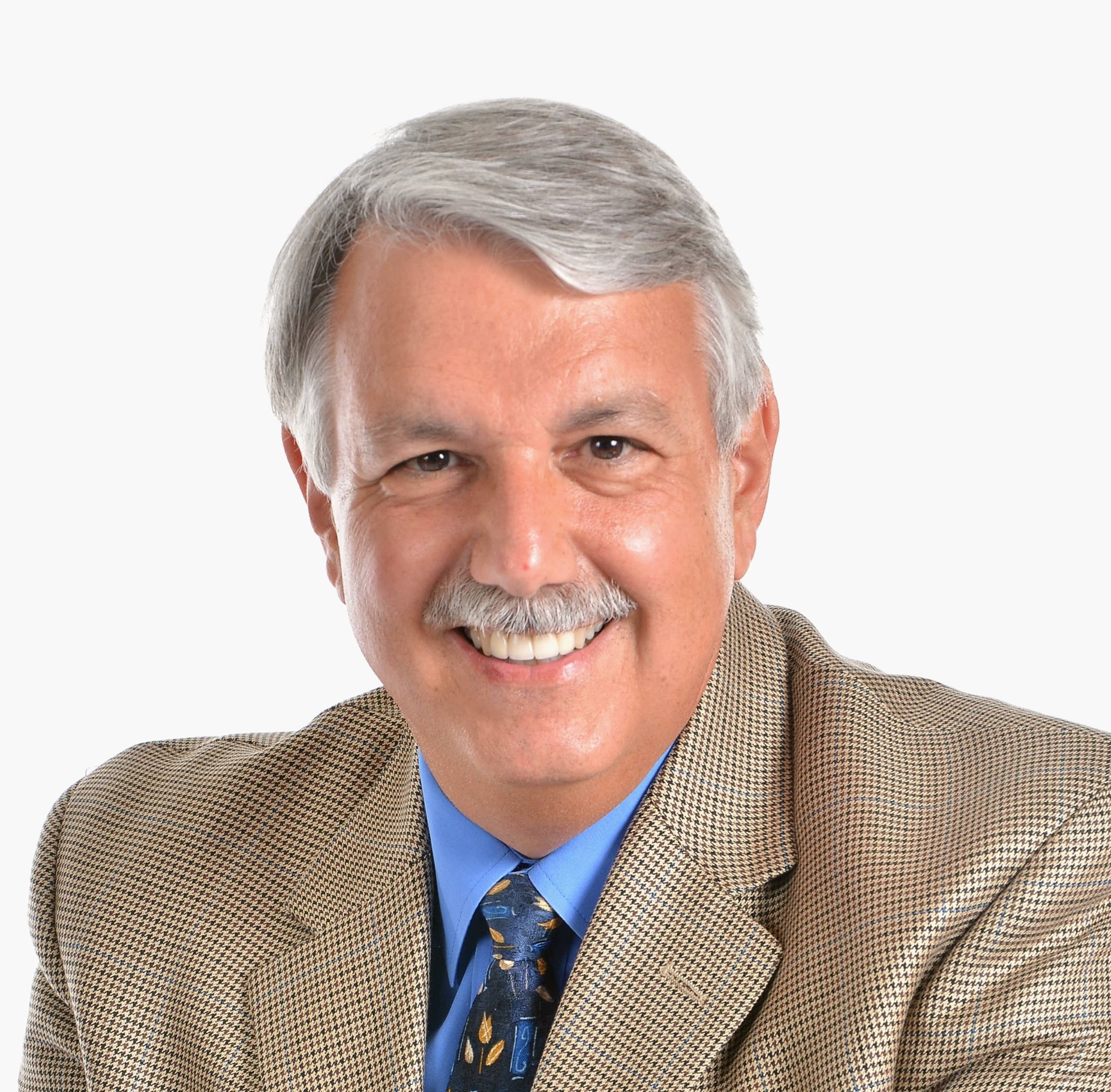 Headshot of an older man with gray hair and a mustache, wearing a tan checkered blazer, blue dress shirt, and dark patterned tie, smiling against a plain white background.