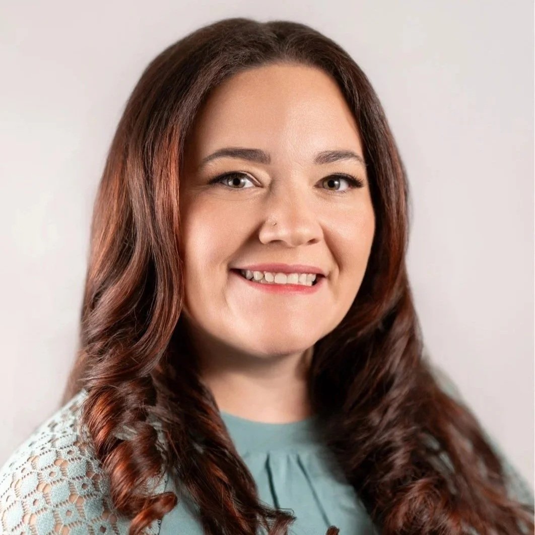 A young woman with long dark hair wearing a beige sweater and black turtleneck holds a disposable coffee cup, standing in front of a plain light background with a green houseplant visible on the right.