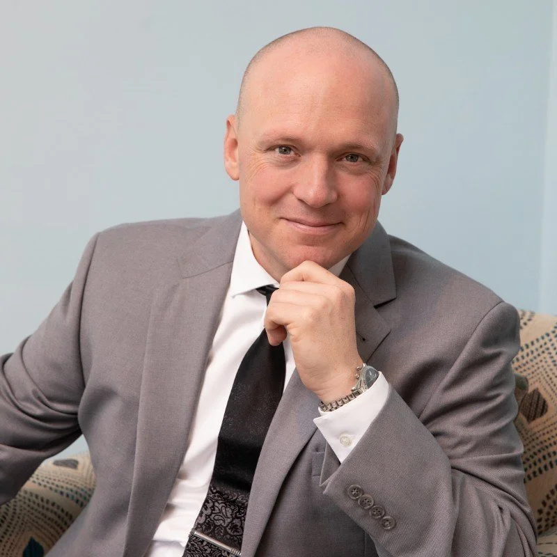 A confident, bald man in a gray suit, white shirt, and black tie, sitting on a patterned chair against a light background, smiling with hand on chin.