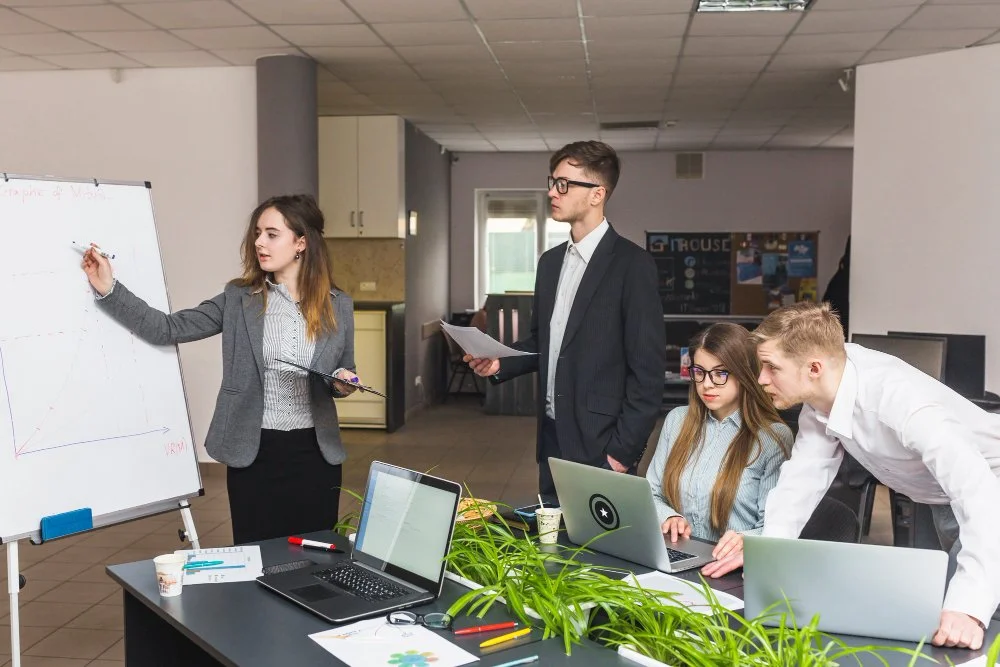 A group of four young professionals in a modern office, engaged in a meeting. One woman stands at a whiteboard presenting, while others are seated and working on laptops or listening attentively.