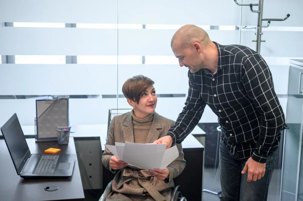 A woman in a wheelchair and a man standing in an office, looking at documents and discussing work.