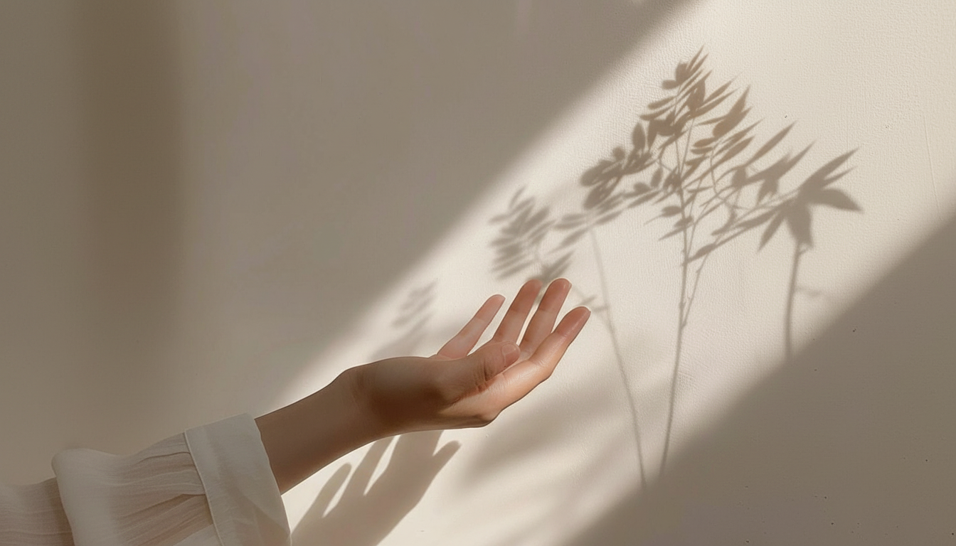 A person's hand reaches out toward a wall with a shadow of plant leaves cast on it, illuminated by sunlight.
