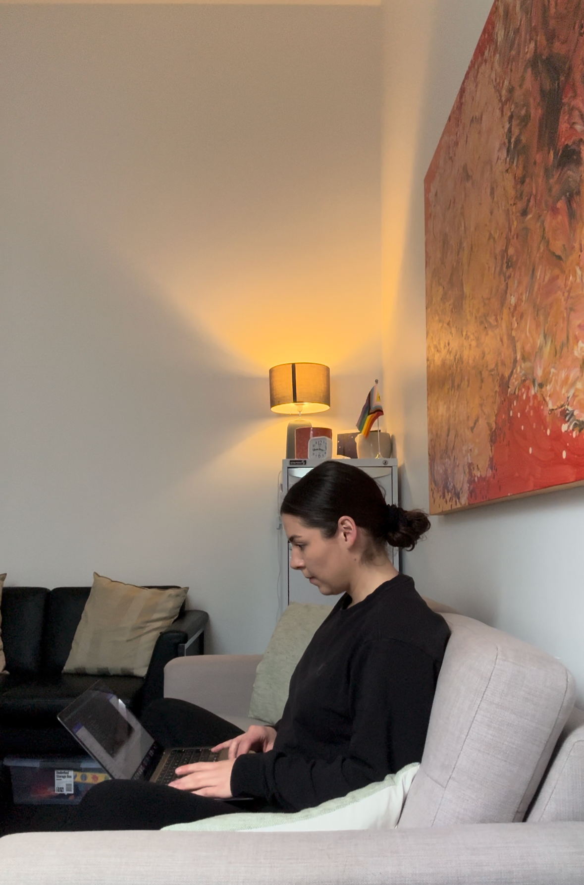 A woman with dark hair tied in a bun sitting on a beige couch, typing on a laptop in a living room with beige and black sofas, a colorful abstract painting on the wall, and a small table with a lamp, a rainbow flag, and some objects in the background