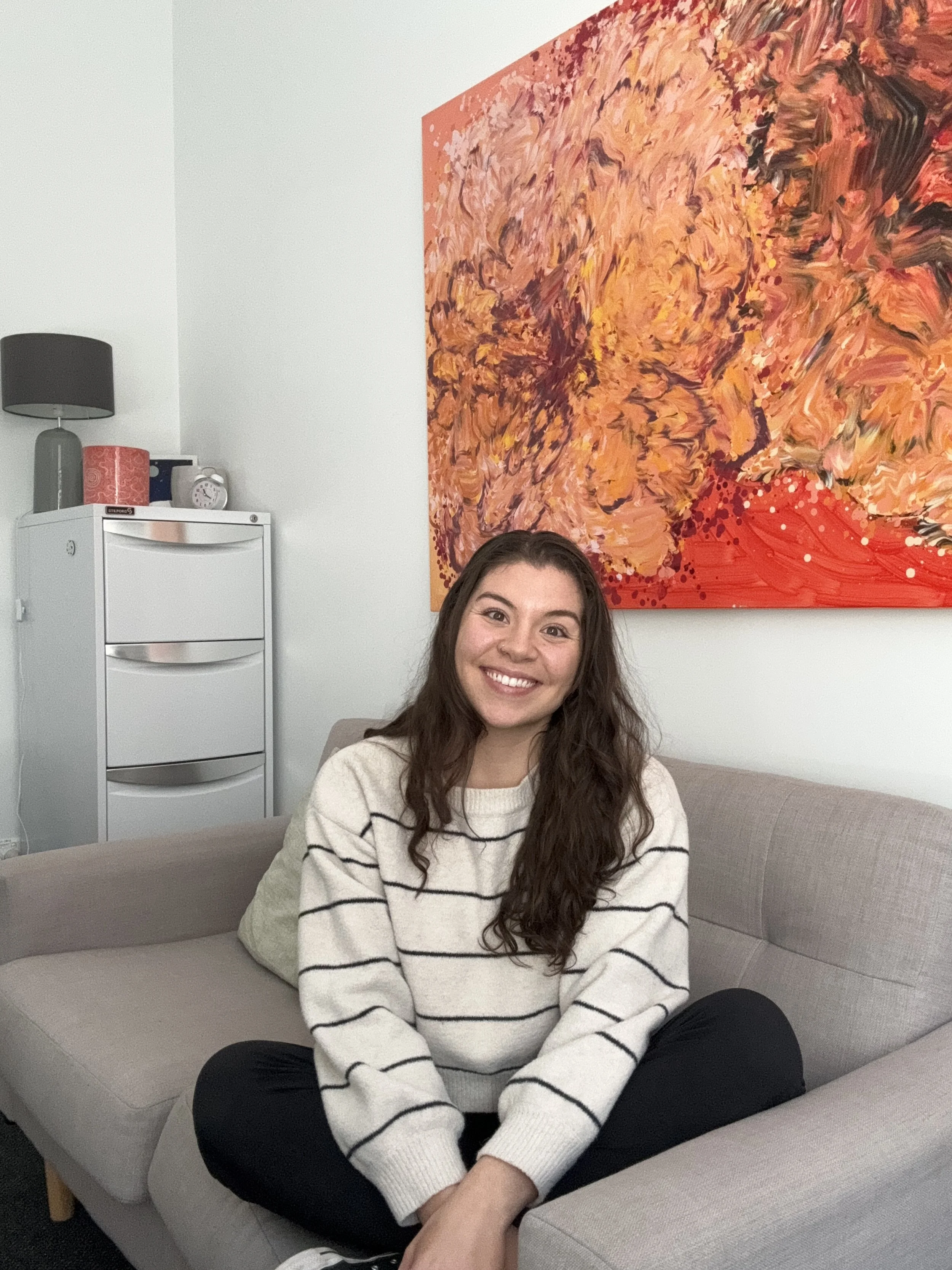 A young woman with long brown hair sitting cross-legged on a beige couch, smiling at the camera, in a room with white walls, a colorful abstract painting behind her, and a white cabinet with a gray lamp and decorative items on top.
