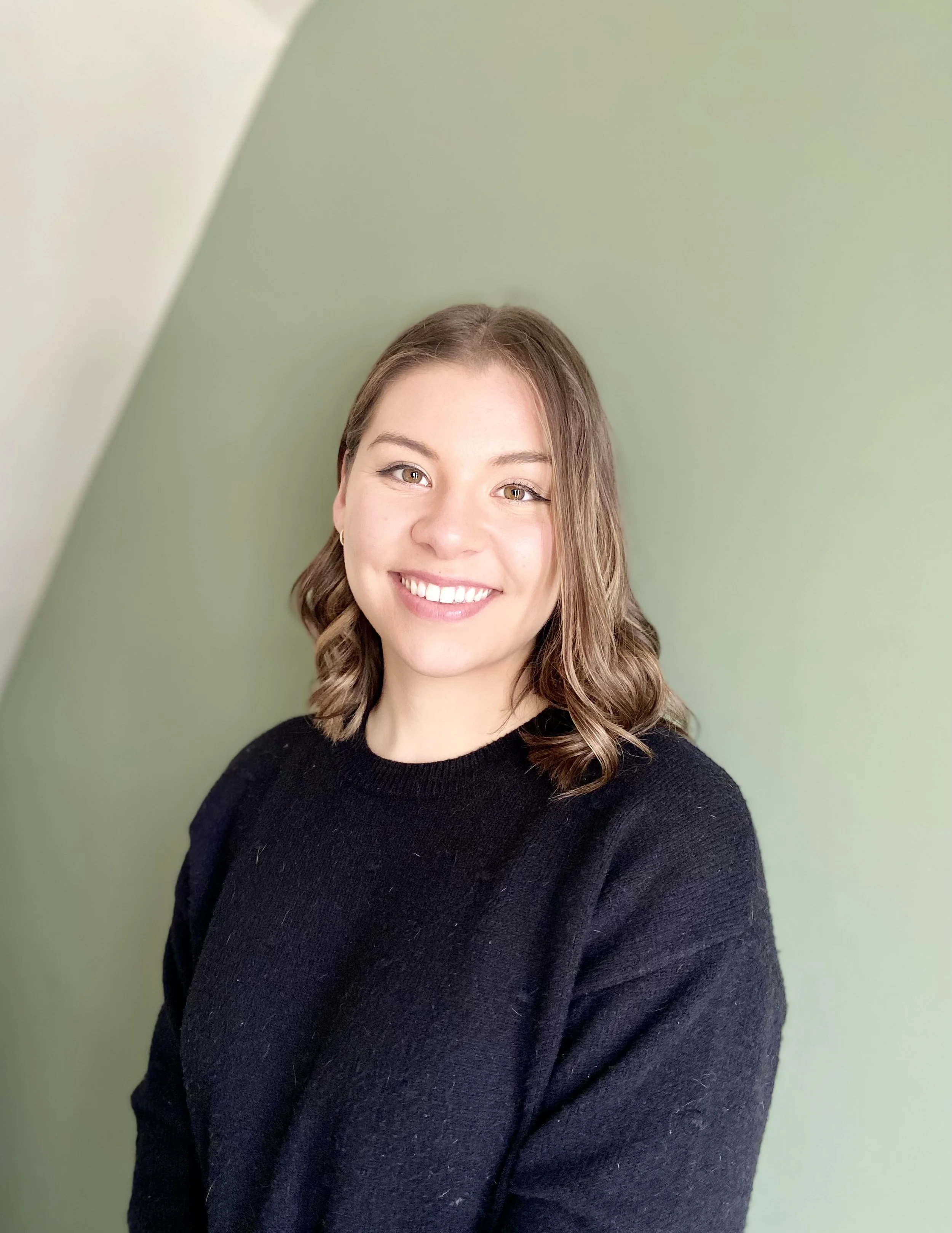 Smiling young woman with shoulder-length wavy brown hair wearing a black sweater against a two-tone green background.
